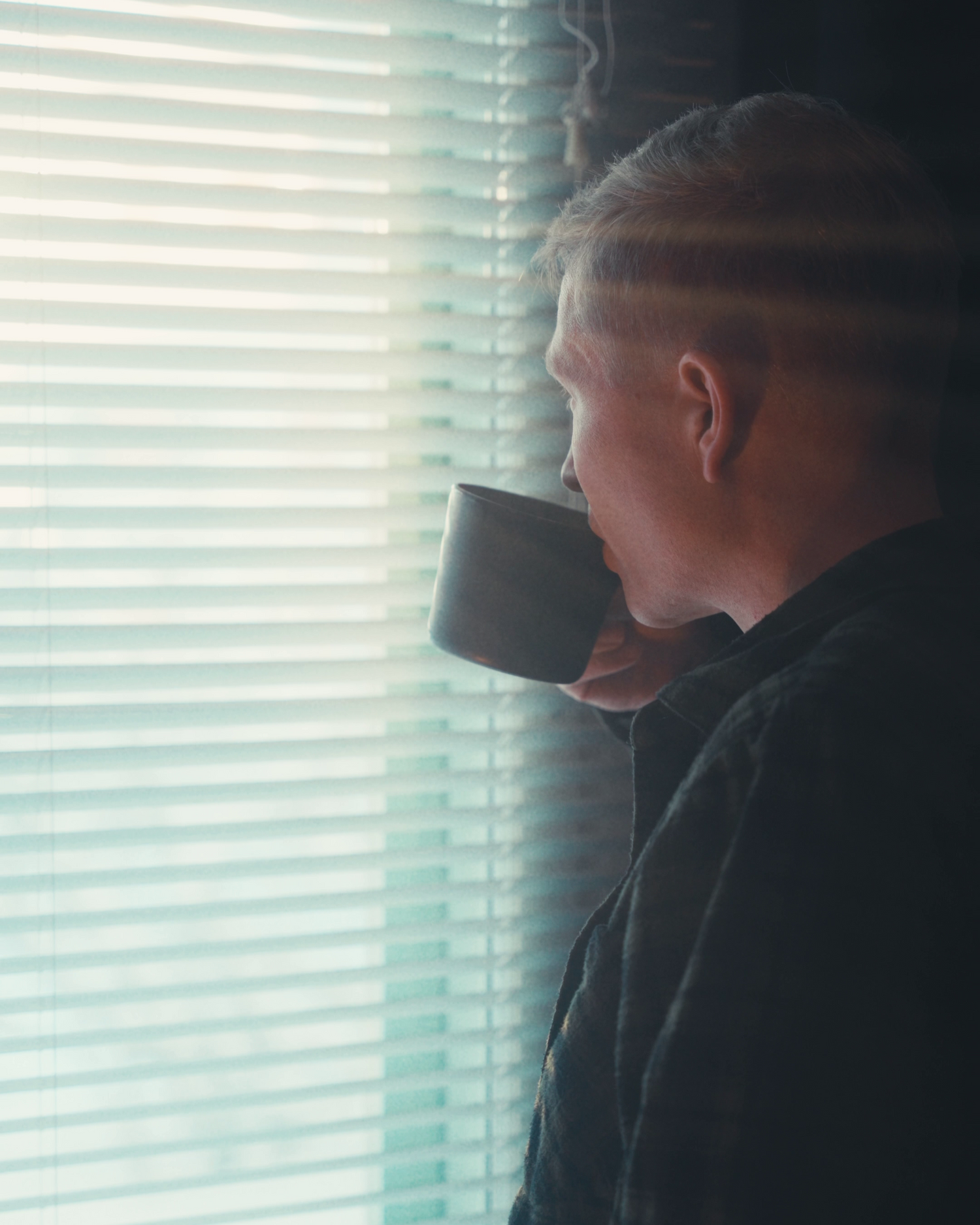 Silhouette near window blinds holding dark mug in moody lighting.