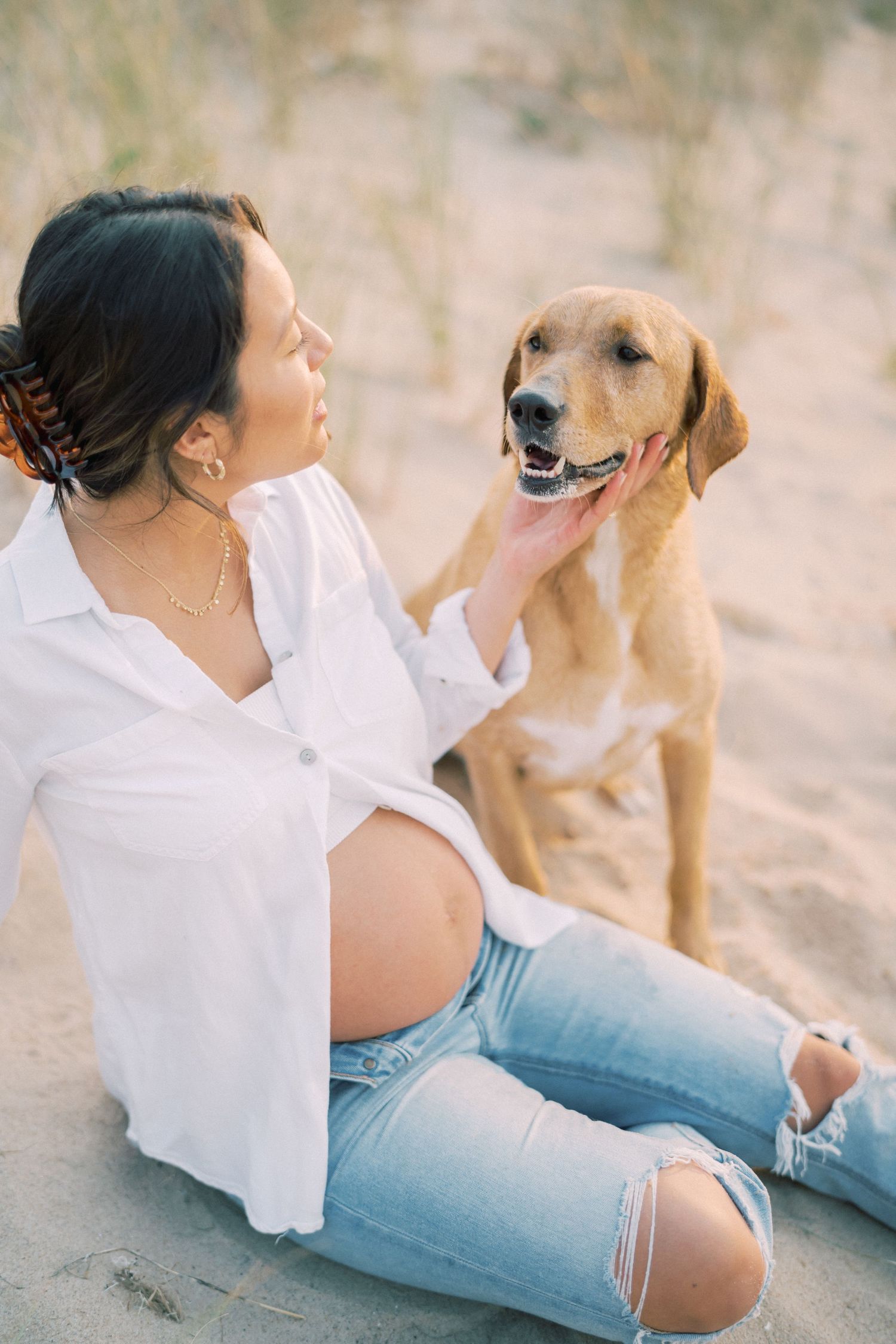 Smiling person in white shirt and ripped jeans sitting on beach with happy golden retriever dog.