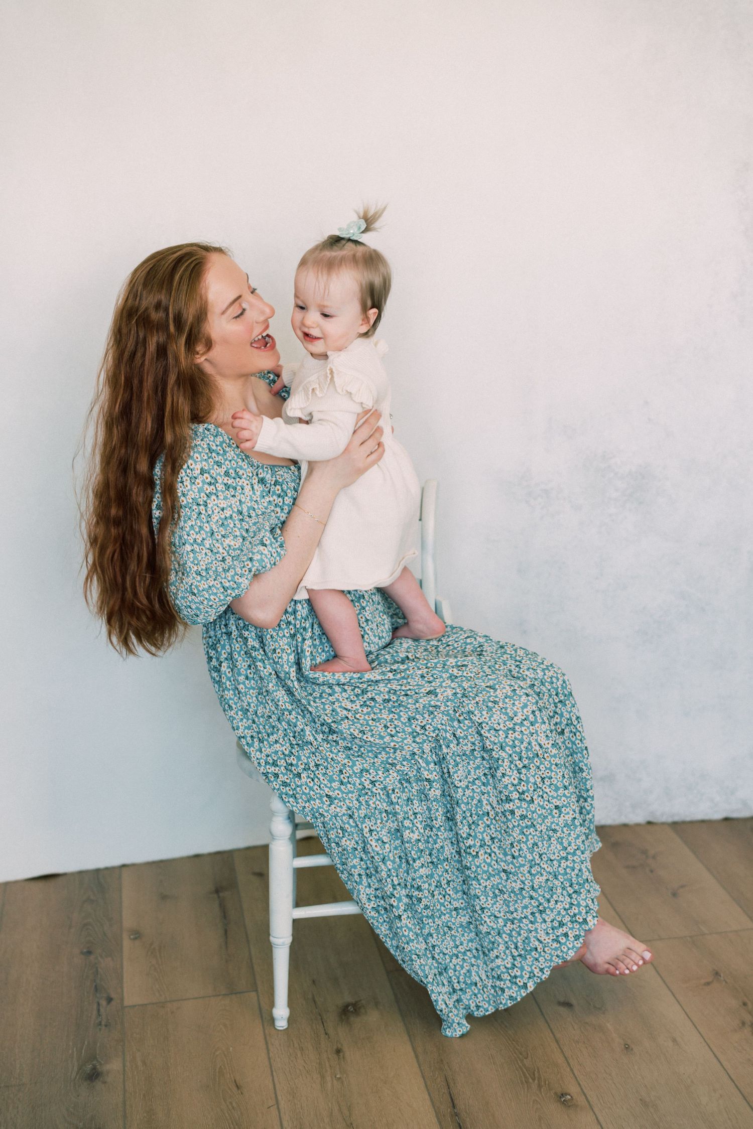 A joyful moment captured between parent and child sharing laughs while sitting on a white chair wearing floral dress.