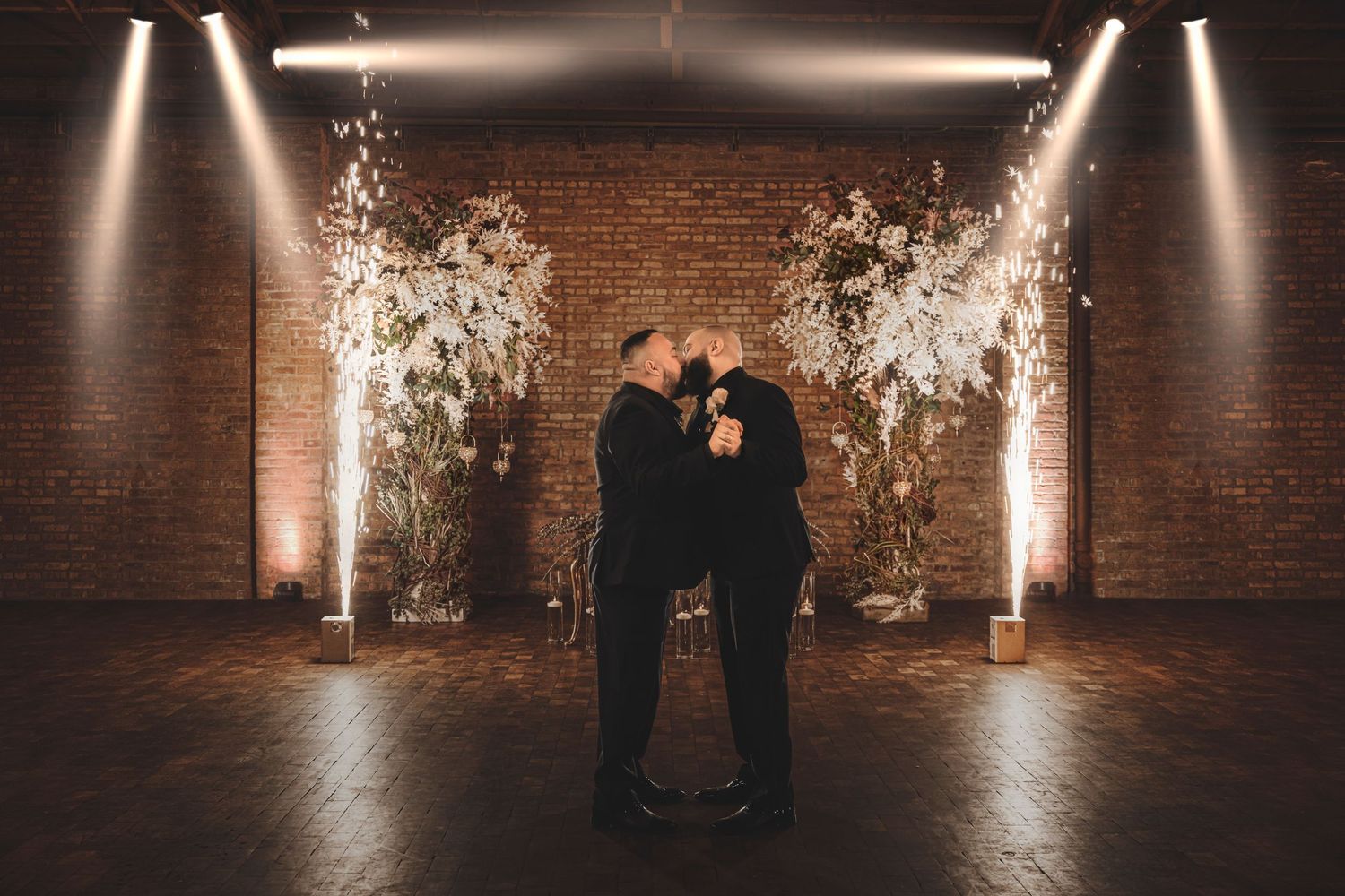 A romantic silhouetted couple stands together beneath florals and spotlights against a rustic brick wall backdrop.
