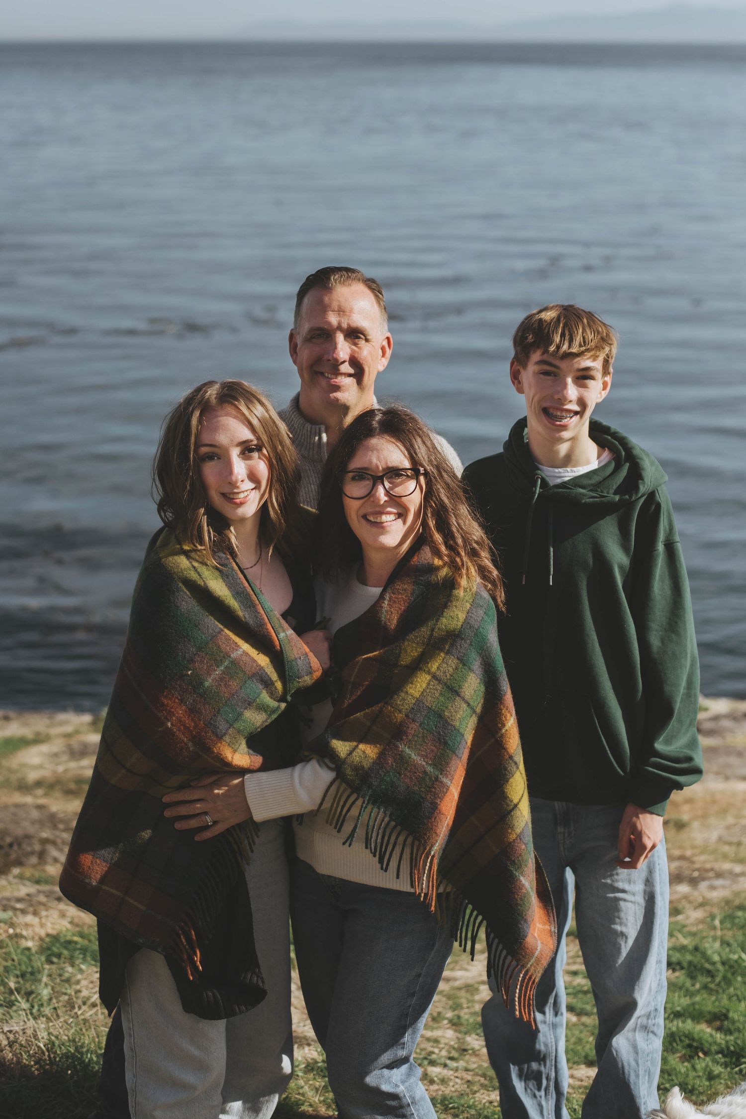 A family of four stands together wrapped in a cozy plaid blanket, smiling warmly on a sunny day by the ocean.