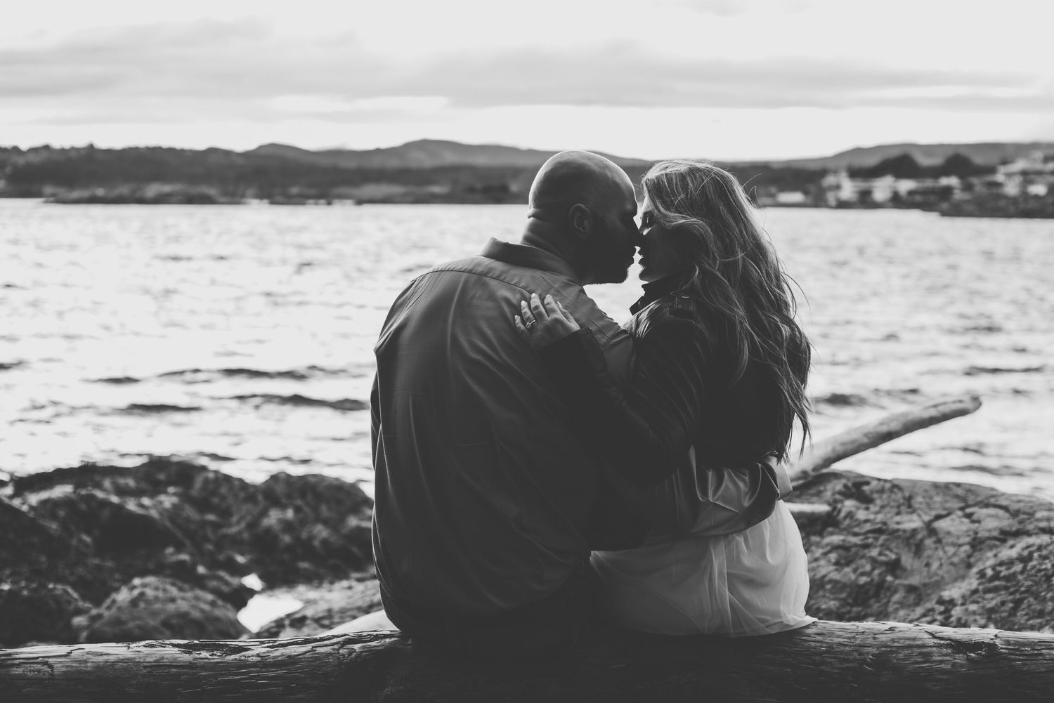 A couple sits closely together on a log by the water, leaning in for a tender moment as they face the serene ocean.