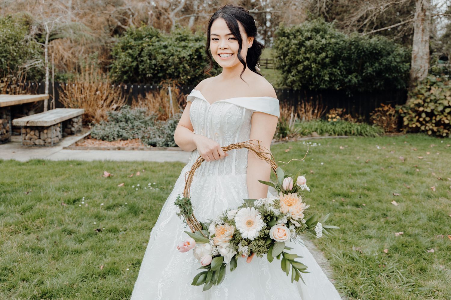 Beautiful off-shoulder white wedding dress with a lush bouquet of cream and blush flowers in a garden setting.