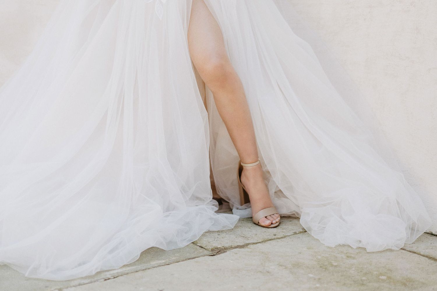 Elegant bridal shoes peek out beneath flowing white tulle wedding dress on stone floor.