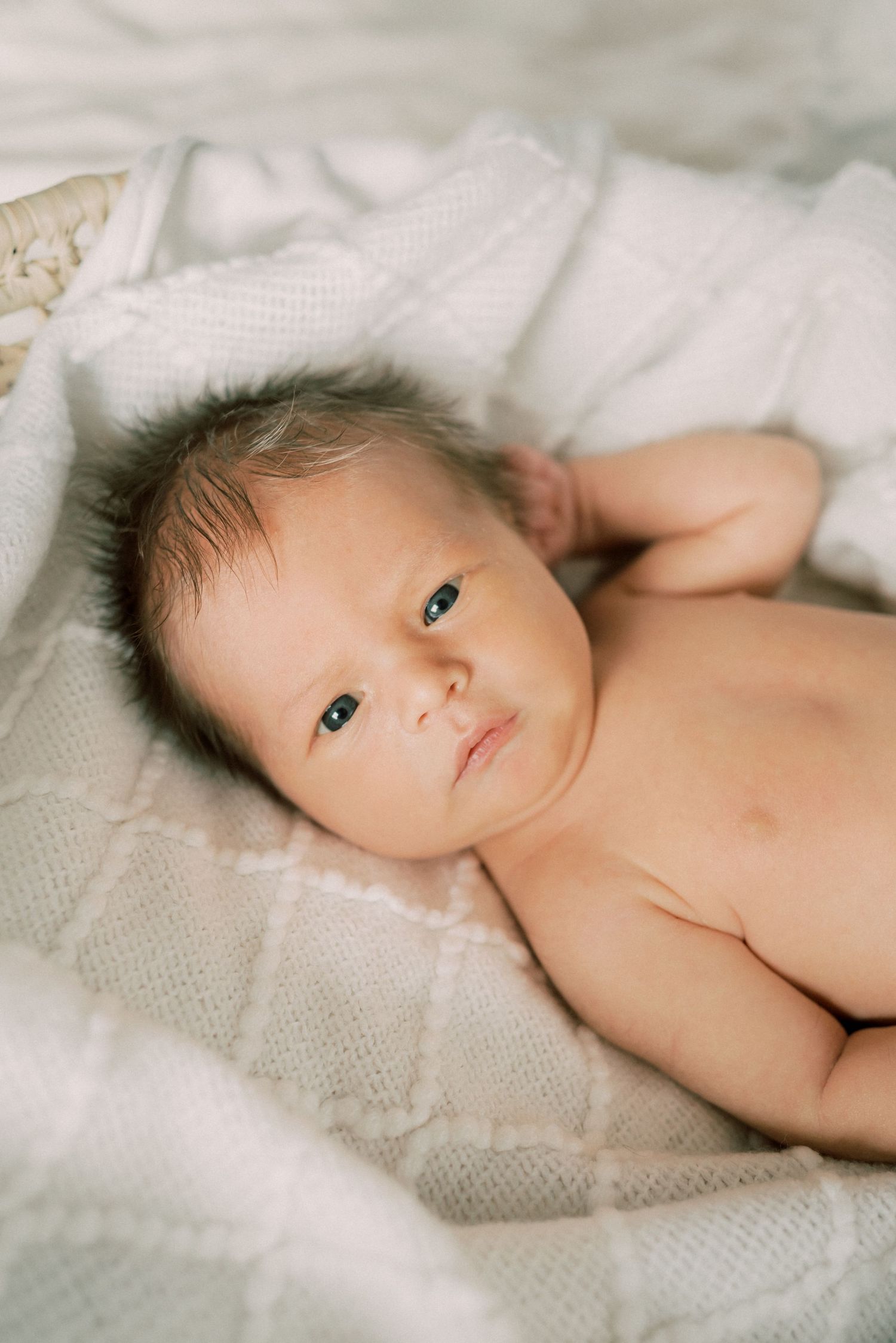A newborn baby lies on soft white textured blankets in a gentle and serene pose.