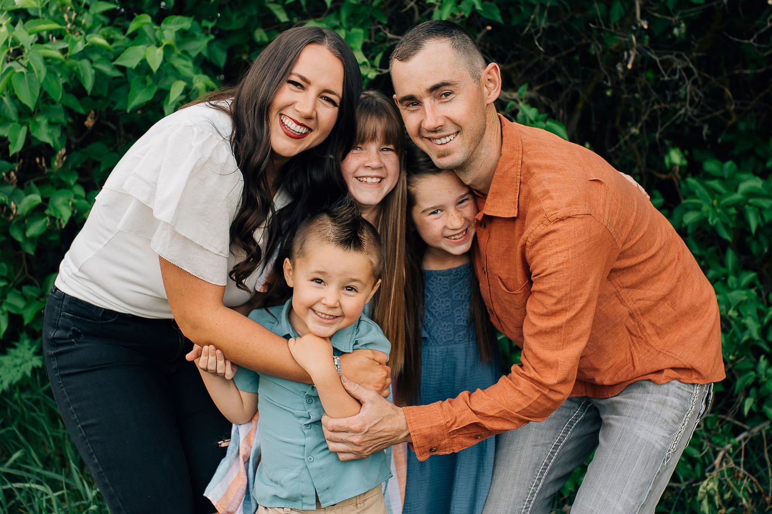 A cheerful family embraces while posing together outdoors against lush green foliage.
