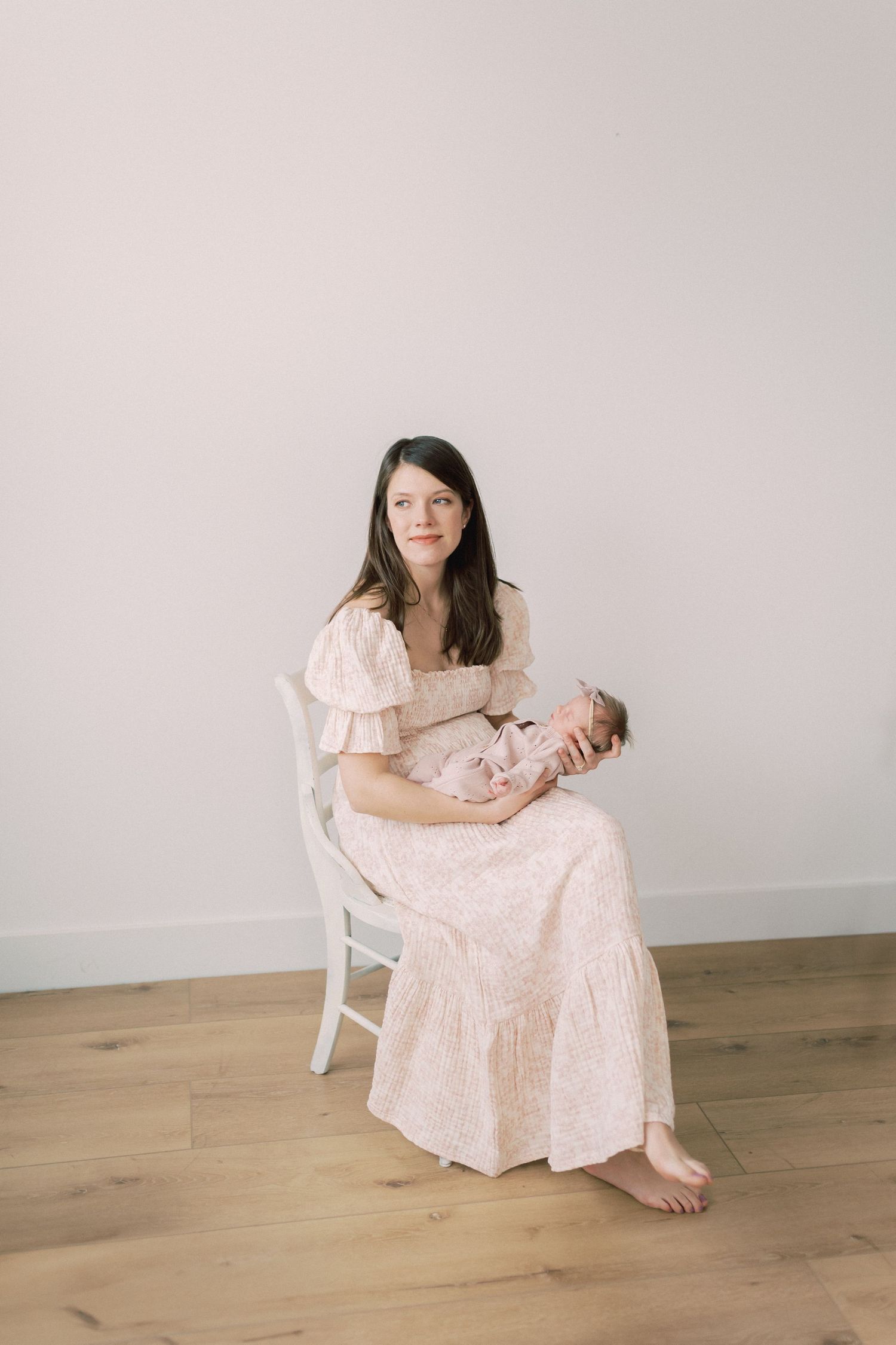 A person in a vintage-style cream lace dress sits on a white chair holding a sleeping newborn baby against neutral background.