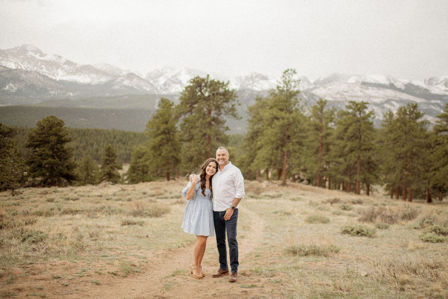 A couple walking together through a scenic mountain meadow in a sepia-toned landscape with evergreen trees.