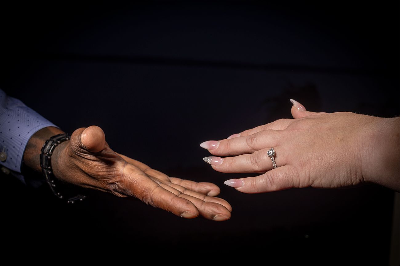 Two hands reaching toward each other against a dark background, one wearing a bracelet and the other a ring.