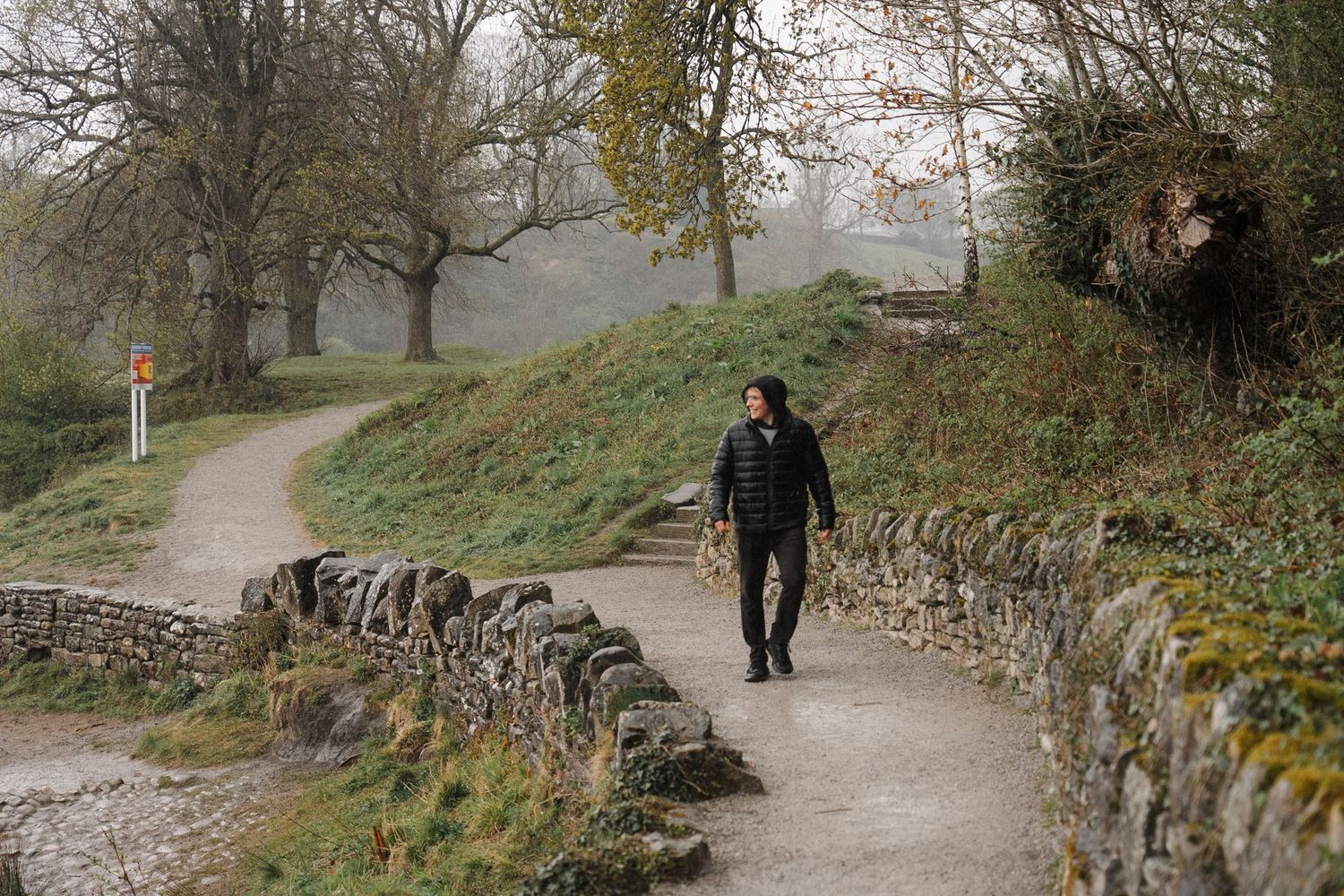 A curved stone-walled pathway leads through a misty autumn forest with a person walking in the distance.