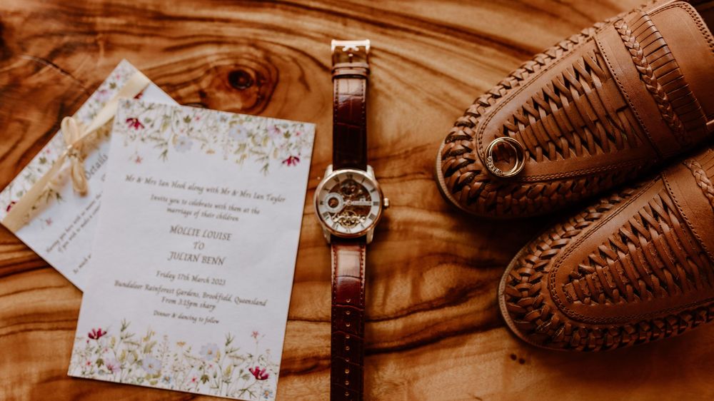 Wedding invitation, brown leather watch, and formal shoes arranged on wooden surface.
