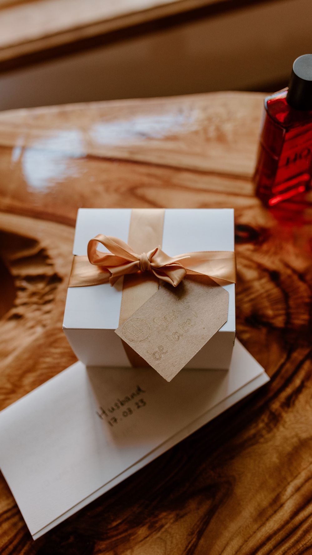 A small white gift box with a tan ribbon bow and heart-shaped tag sits on a wooden surface next to a red bottle.