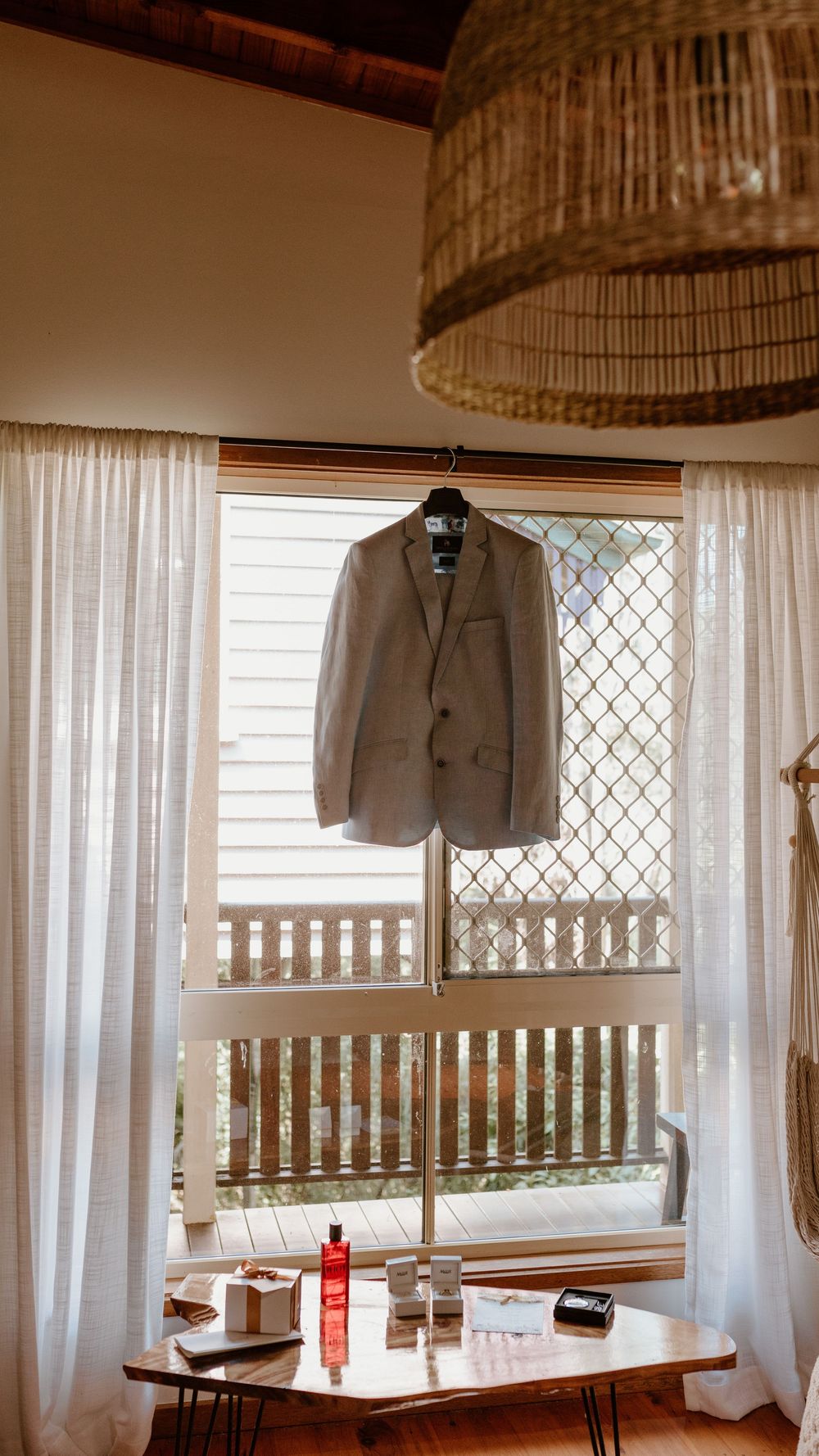 Light gray suit jacket hanging by window with woven lamp shade and white curtains.