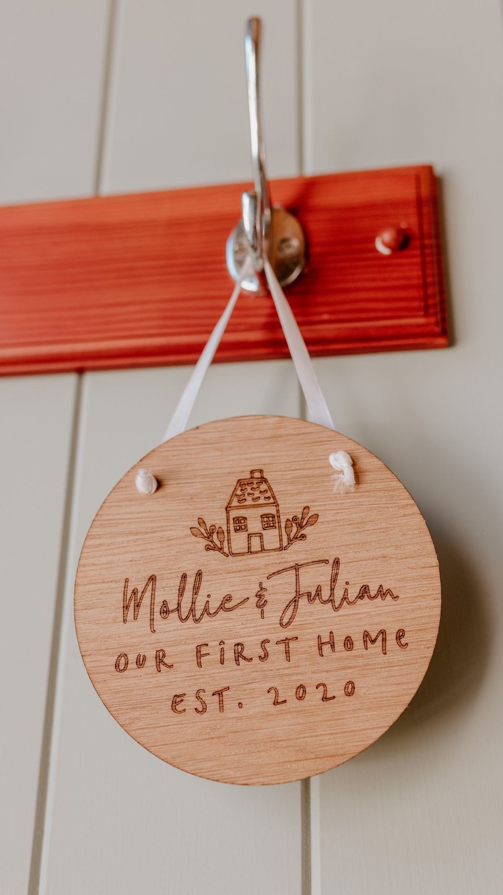 A wooden circular plaque engraved with 'Mollie & Julian Our First Home Est. 2020' and a small house design hangs from a hook.