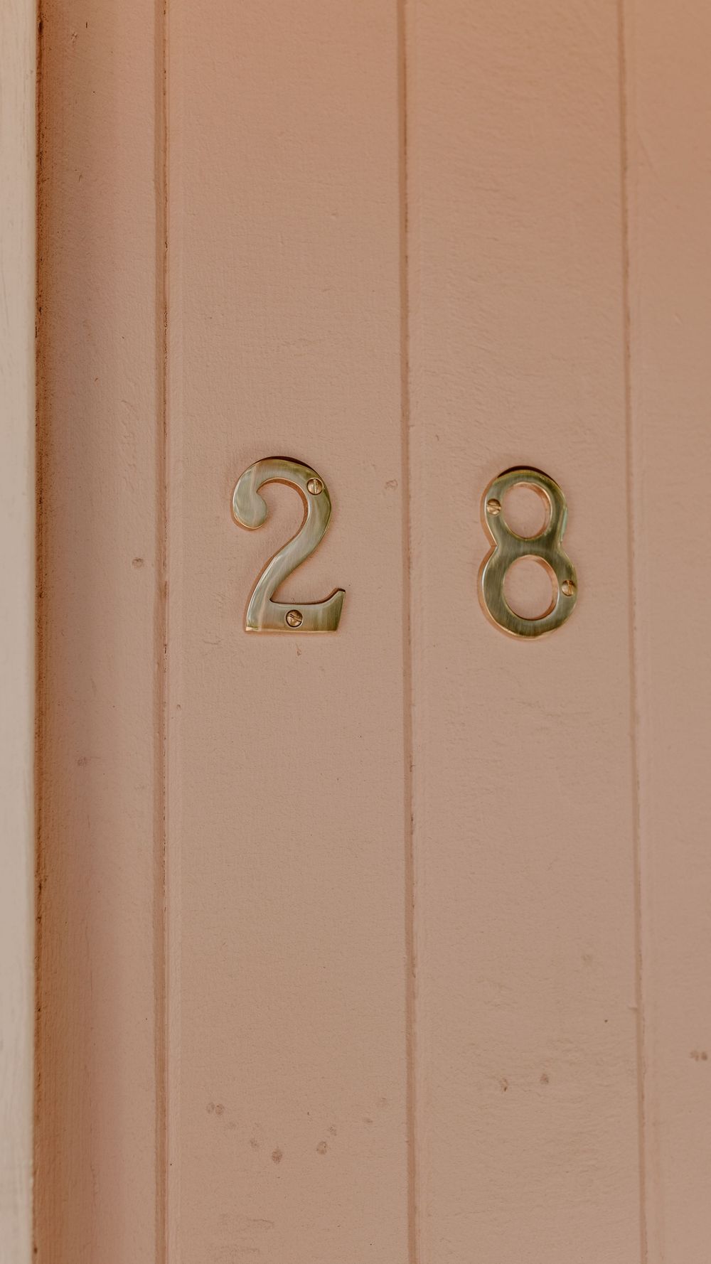 Gold house numbers 28 mounted on a beige wooden wall.