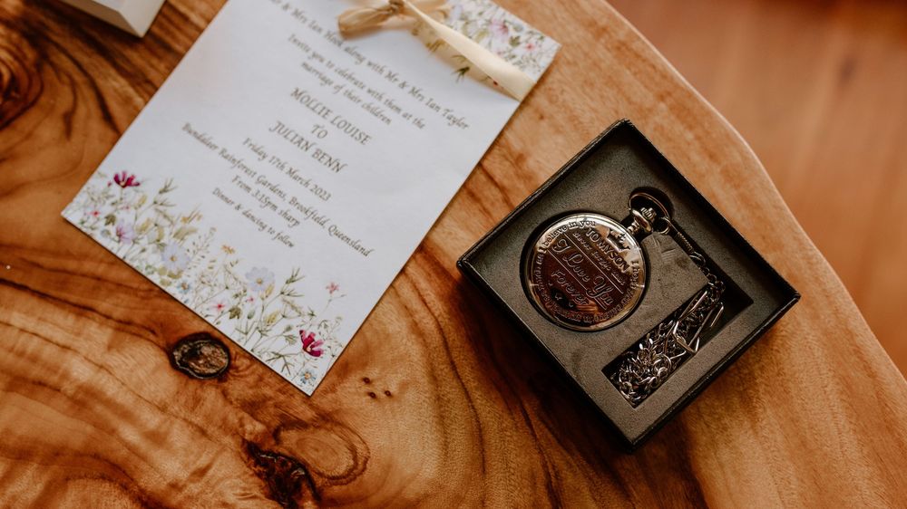 A floral wedding invitation card displayed next to a vintage ring box on peach-colored fabric.