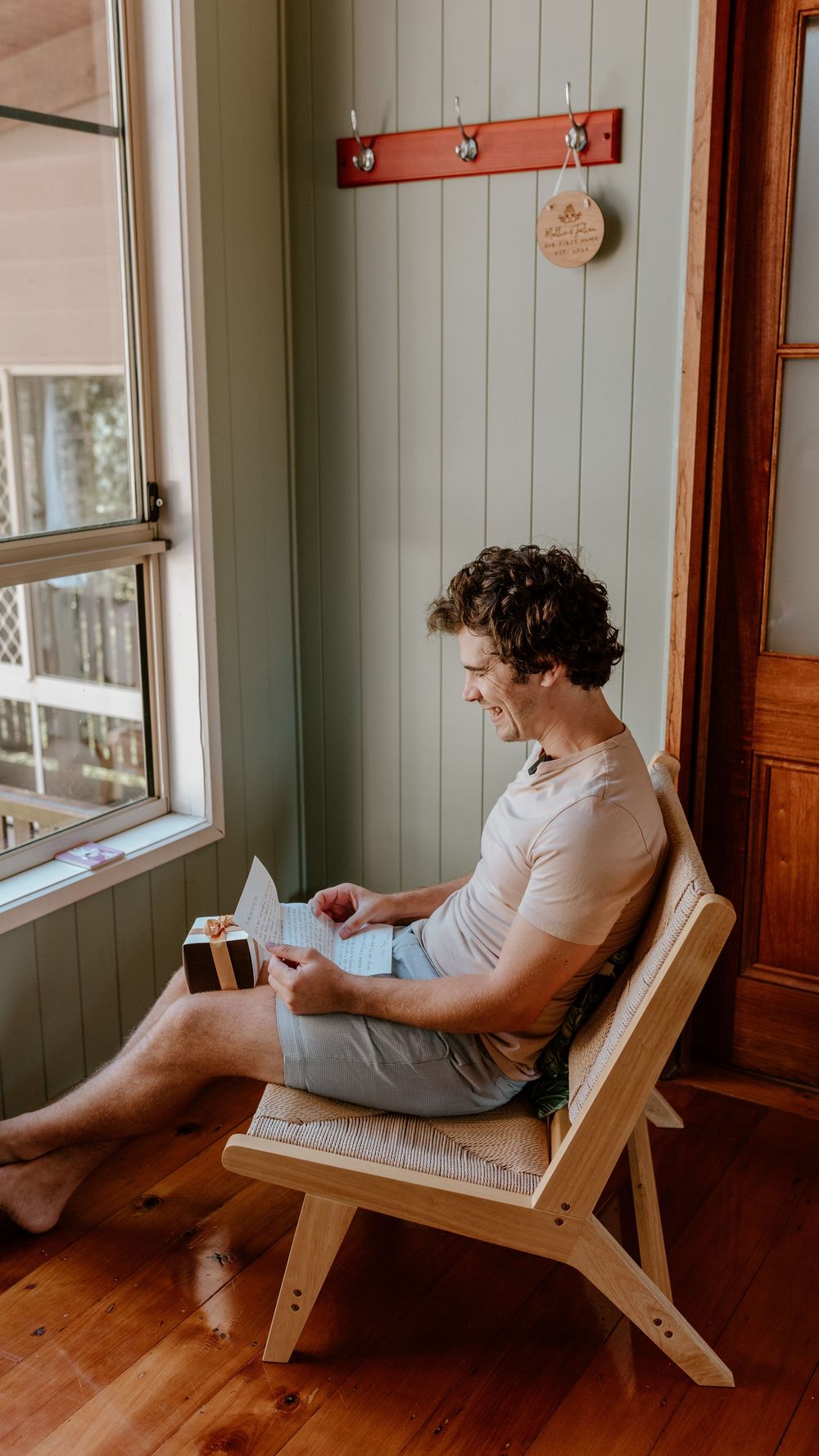 A person in a white shirt sits reading in a wooden lounge chair by a window in a rustic interior.
