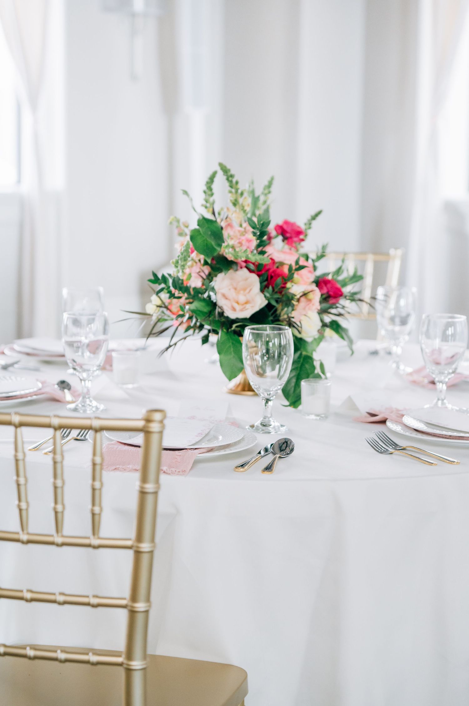 Elegant table setting with pink and peach floral centerpiece, white linens, crystal glassware and gold chiavari chair.