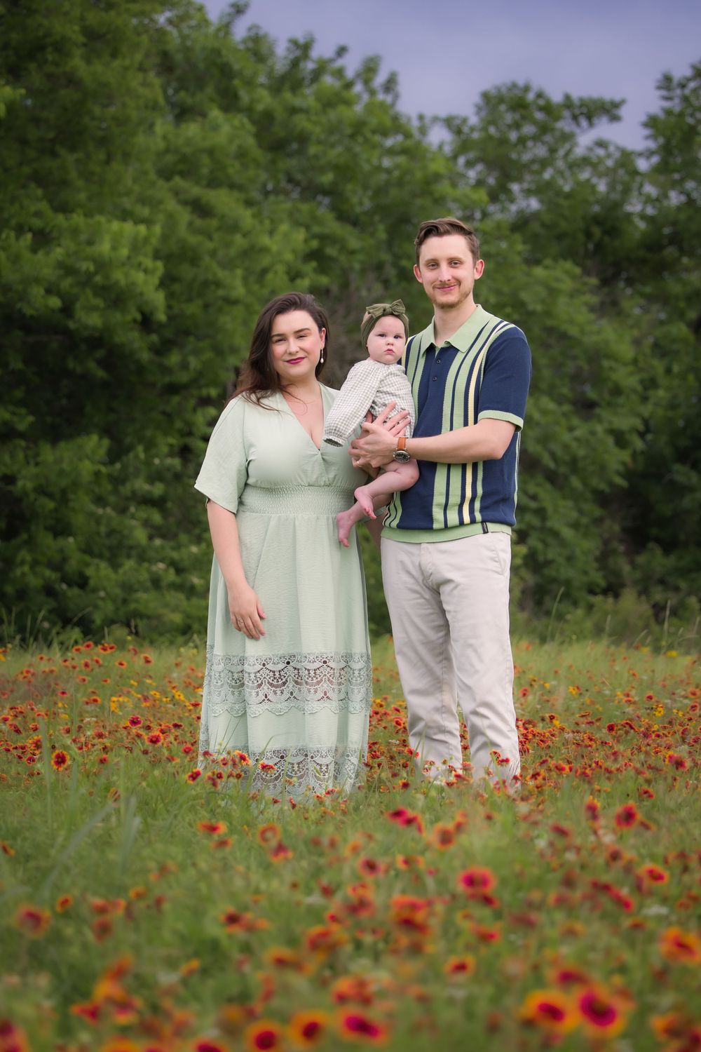 Family standing together in a vibrant red wildflower field with lush green trees in the background at sunset.