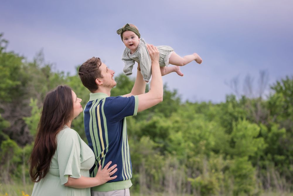 A person lifts a toddler high in the air while another person watches outdoors against a green forest background.