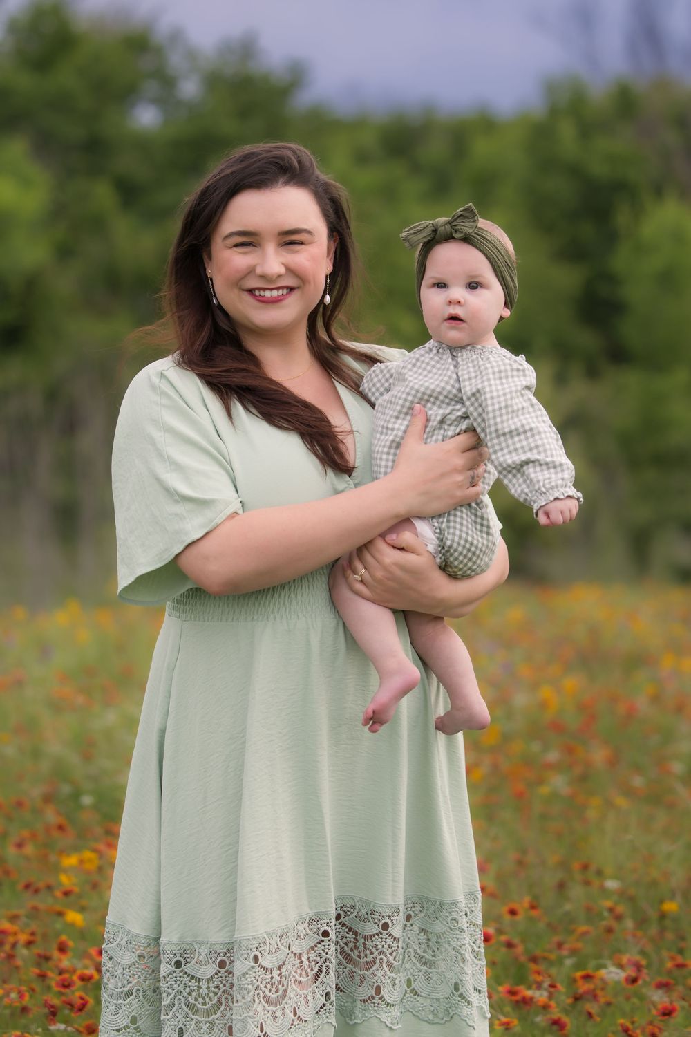 Mother and baby pose together in a sage green dress and gingham outfit amidst a vibrant wildflower field.