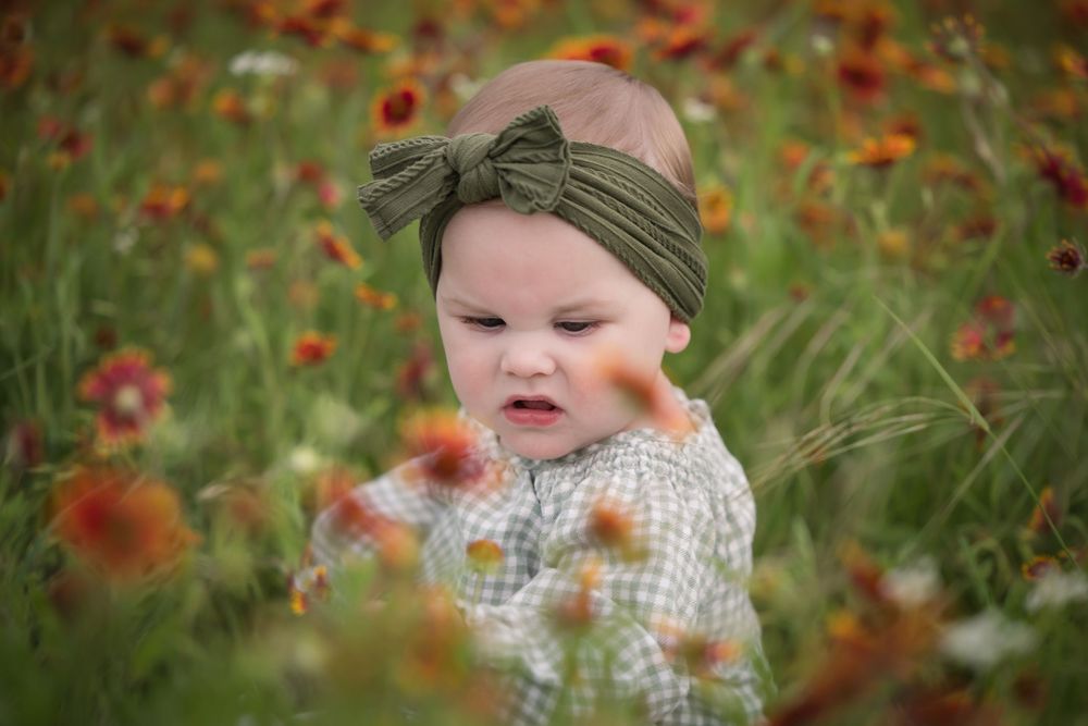 Baby wearing a headband sits among orange wildflowers in a spring meadow.