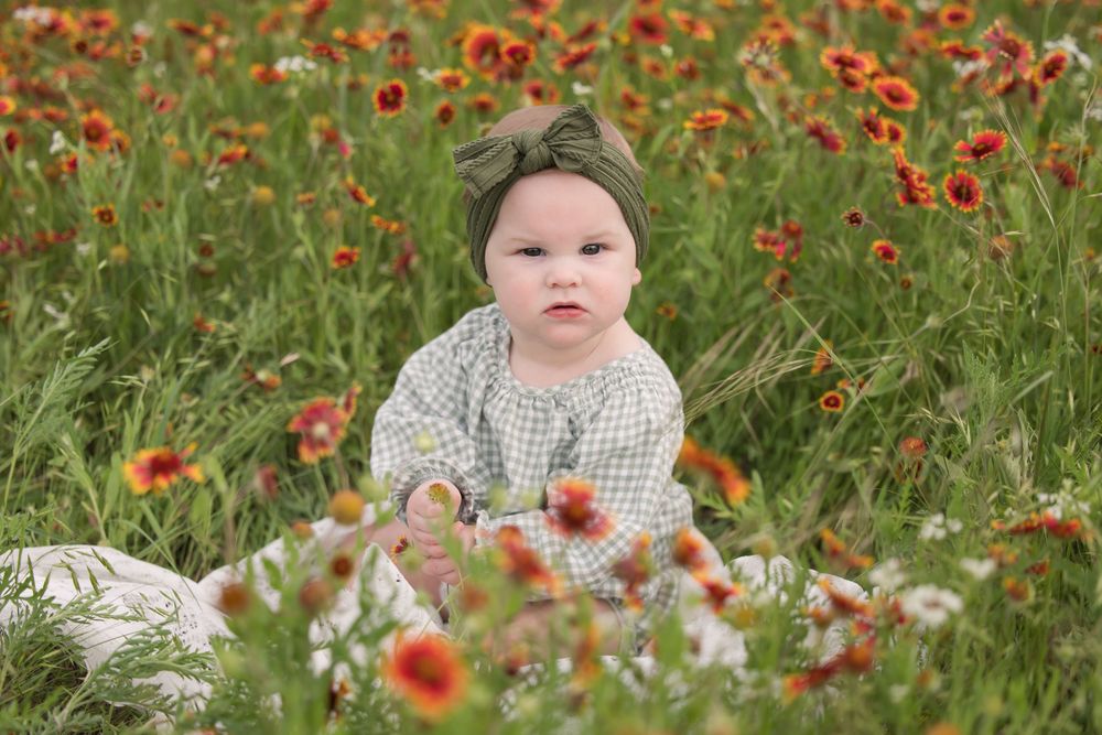 A baby sits among beautiful orange wildflowers in a lush green field wearing a patterned outfit.