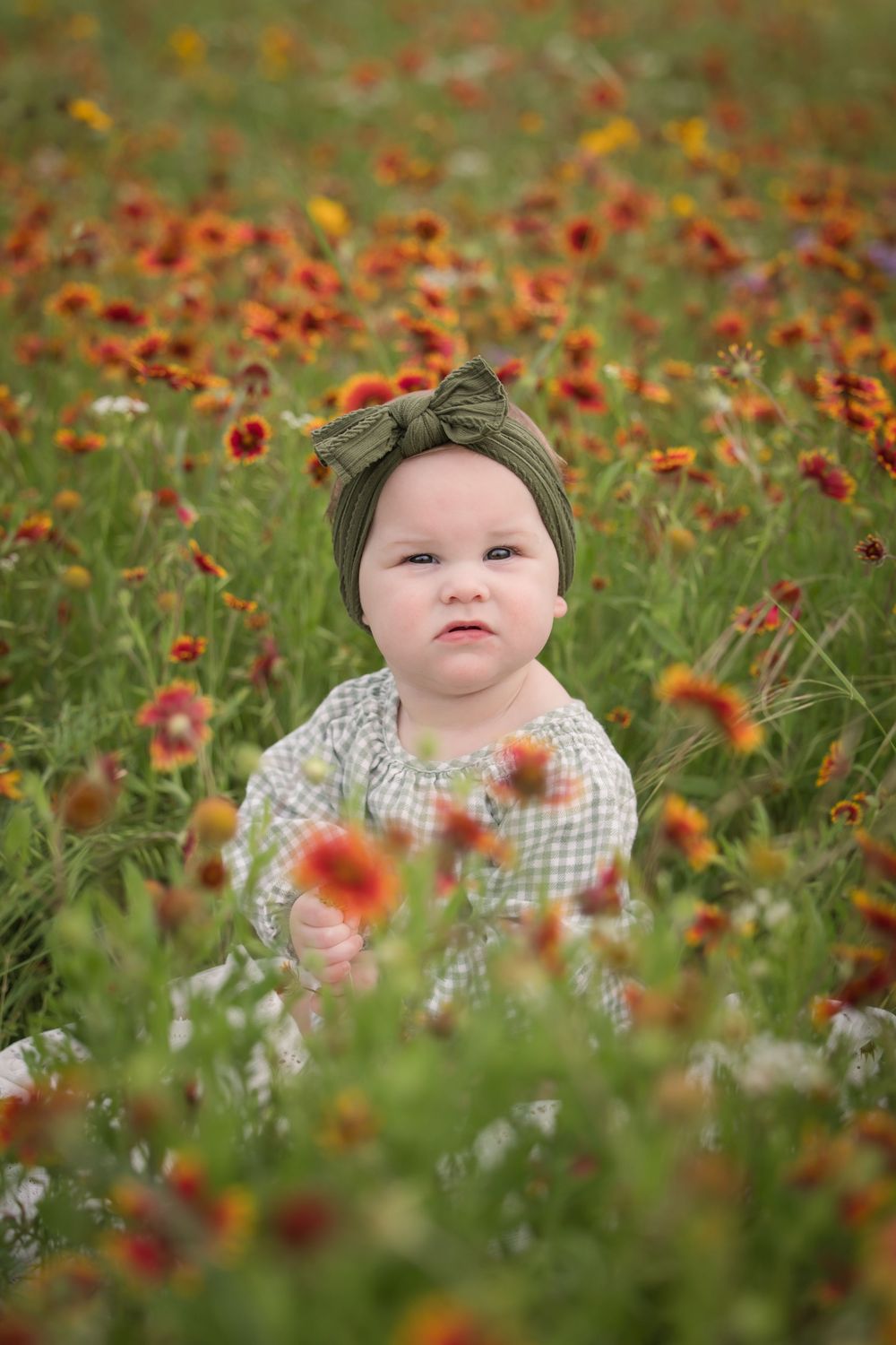 A small child wearing a green headband and gingham dress sits among blooming orange wildflowers in a meadow.