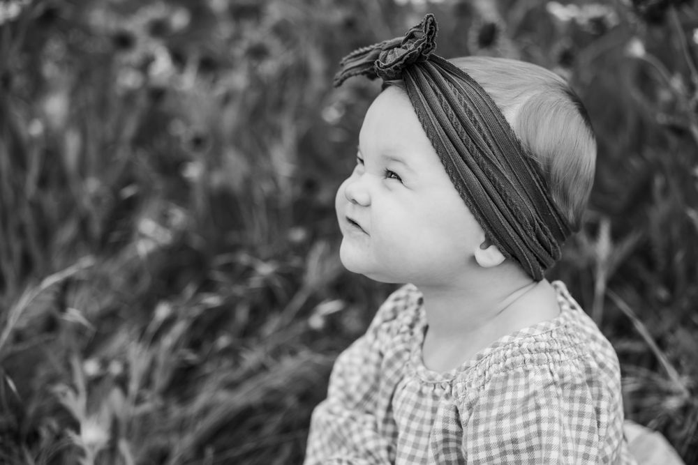 A black and white portrait of a toddler in gingham dress and headband looking upward thoughtfully.