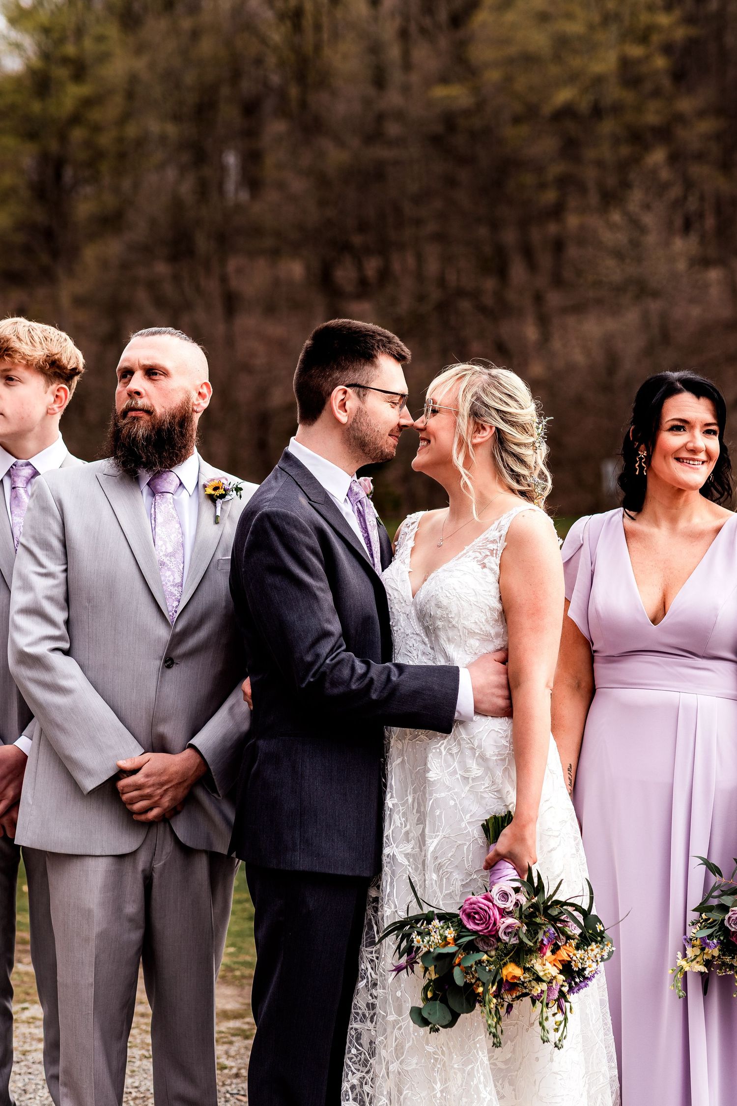 Wedding party members gather outdoors in formal attire with purple bridesmaid dresses and a pink floral bouquet.