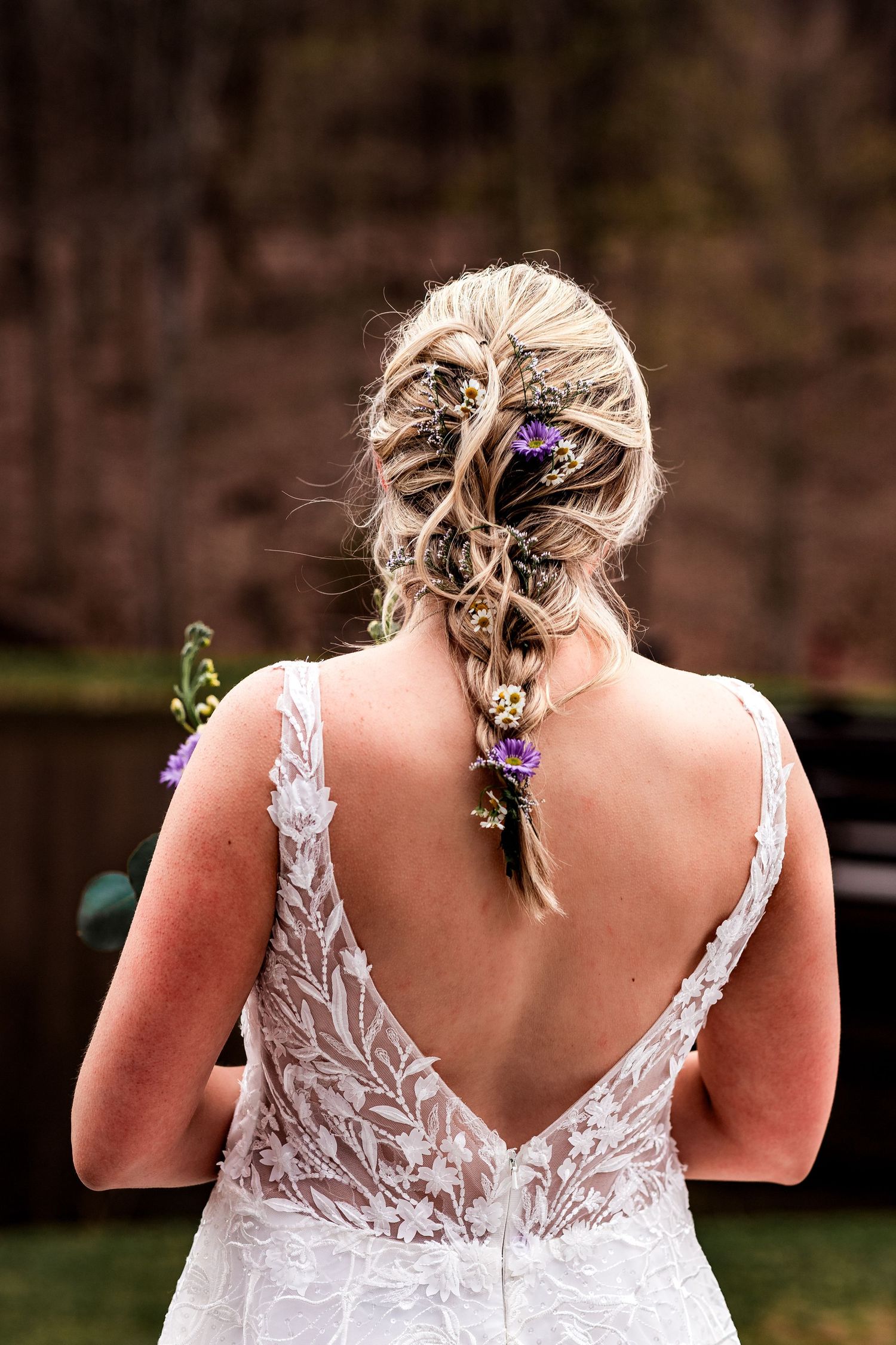 Back view of bride in lace wedding dress with blonde braided hairstyle adorned with small purple flowers.