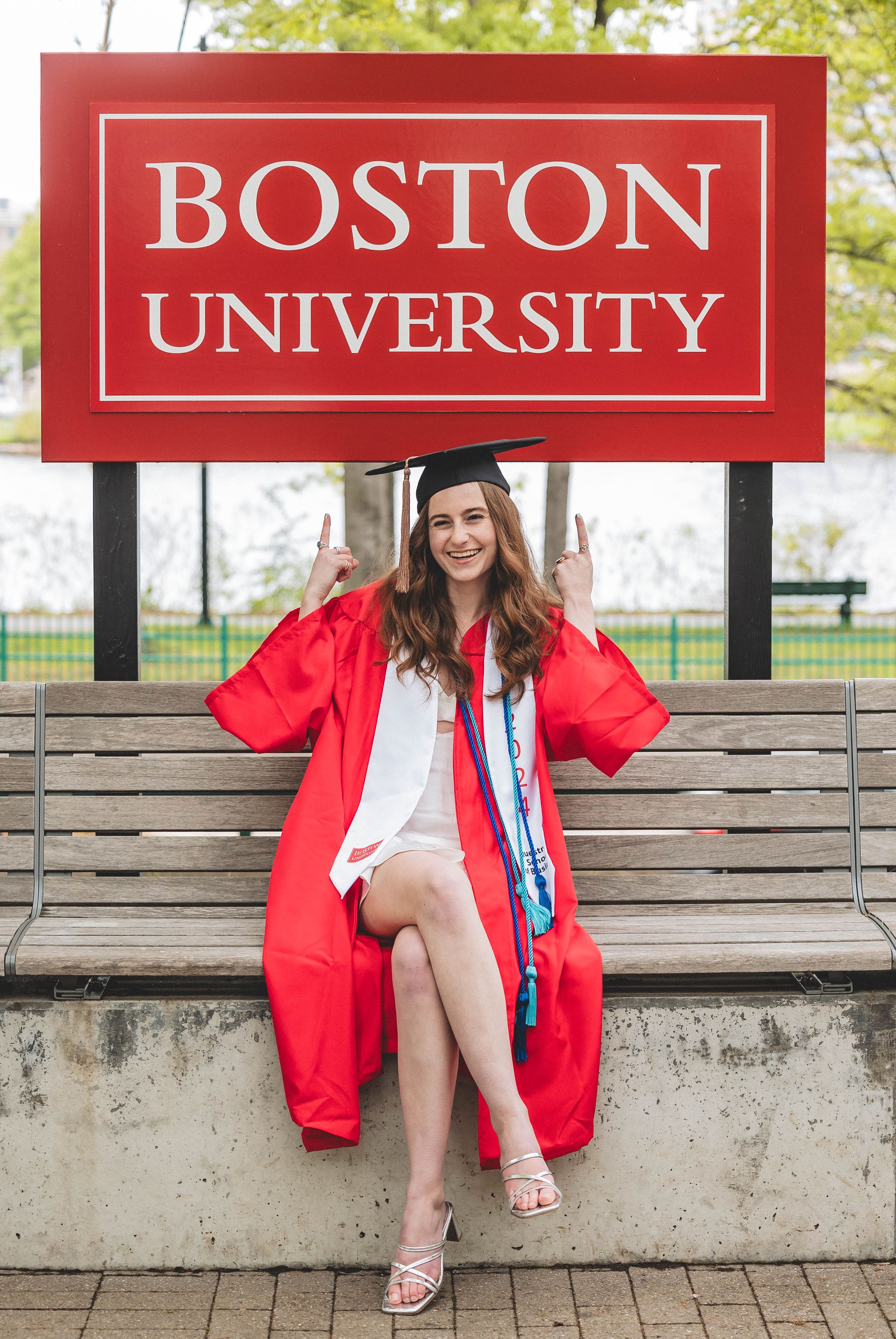 A graduate in red academic regalia sits on a bench beneath a Boston University sign during commencement.