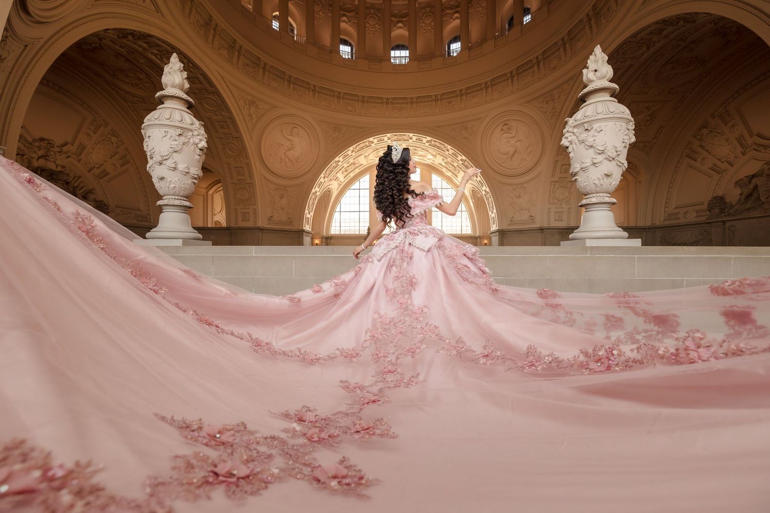 A quinceanera stands in an ornate marble rotunda with a dramatic flowing pink dress train cascading down the stairs.
