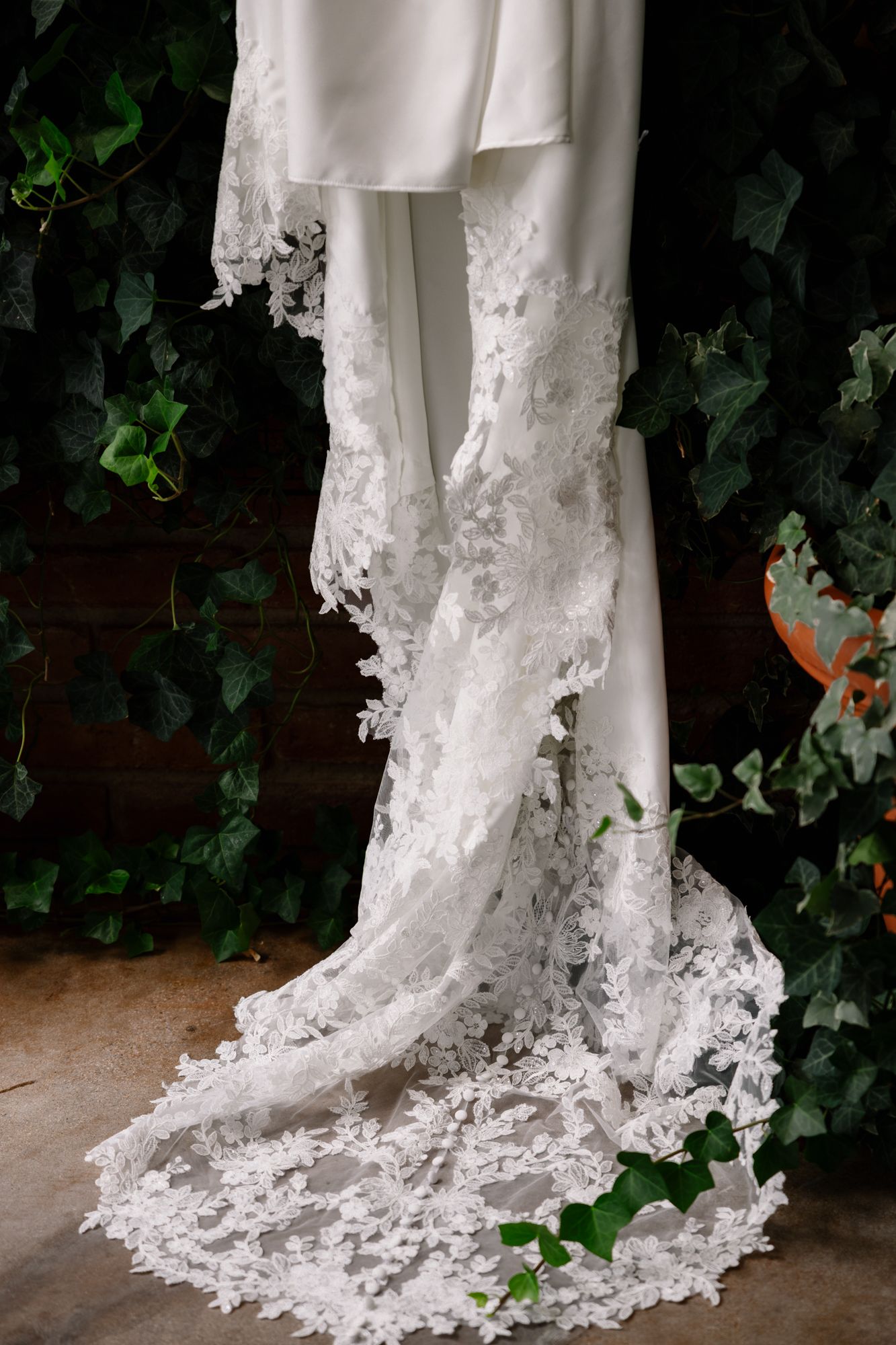 Elegant white wedding dress train with intricate lace detailing hangs against ivy-covered brick wall background.