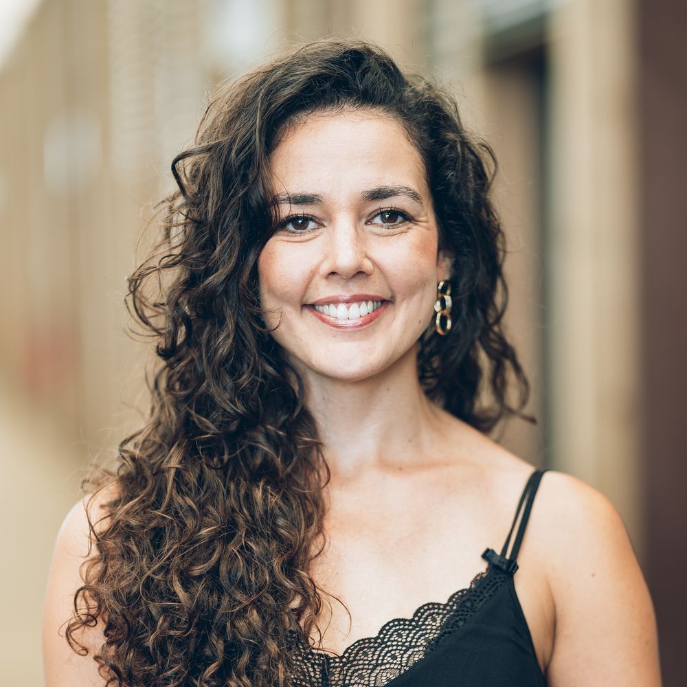 Woman with dark curly hair wearing black lace top smiles warmly in professional beauty portrait series.