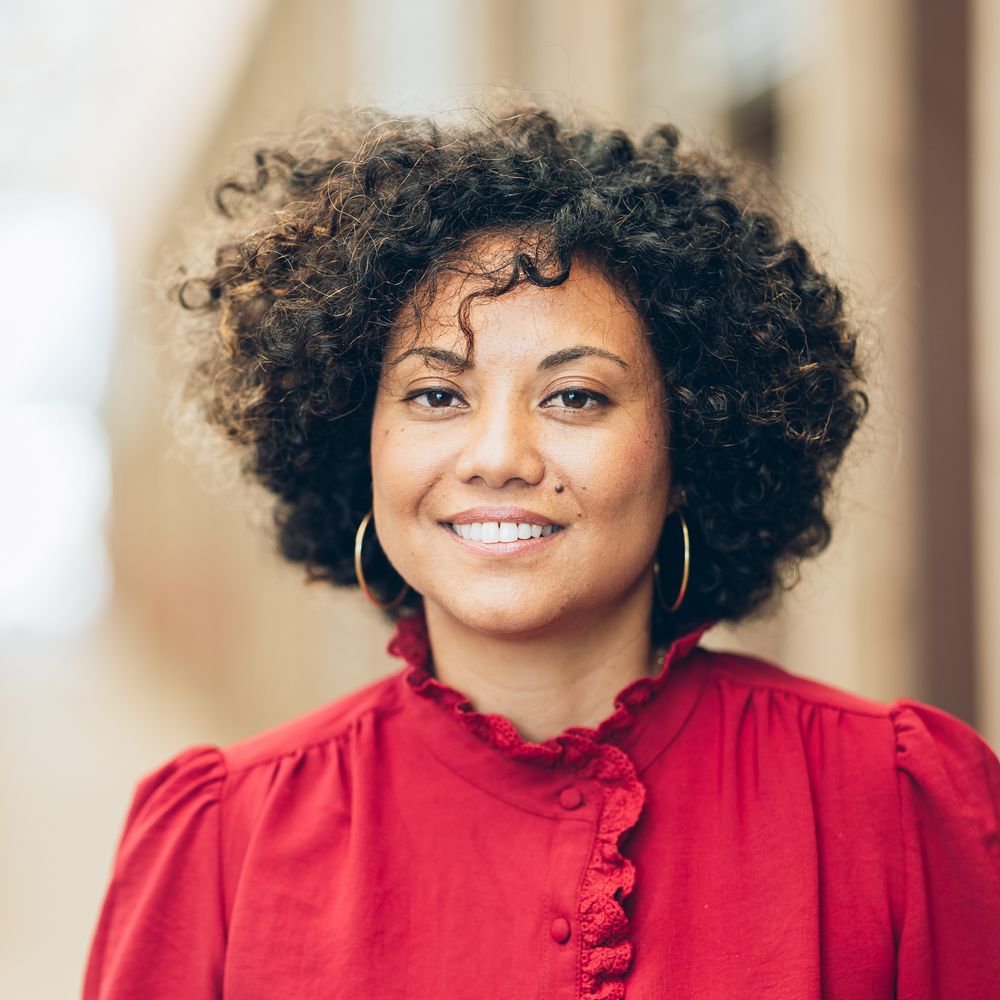 Person with natural curly hair wearing a bright red ruffled blouse and gold hoop earrings poses against light background.