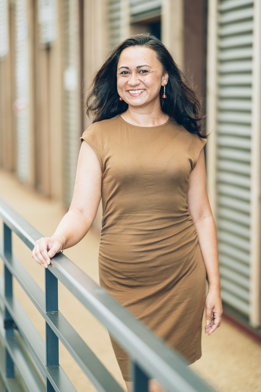 Person wearing brown sleeveless dress poses on staircase while smiling and looking away from camera.
