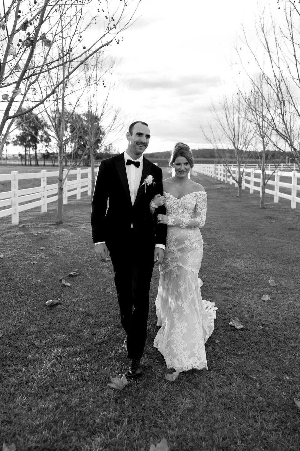A couple in formal wedding attire walks along a path with white fencing on a cloudy day.