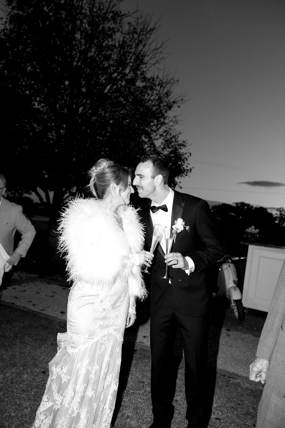 Elegant black and white wedding photo showing couple in formal attire including bride in lace gown with feather wrap at night.