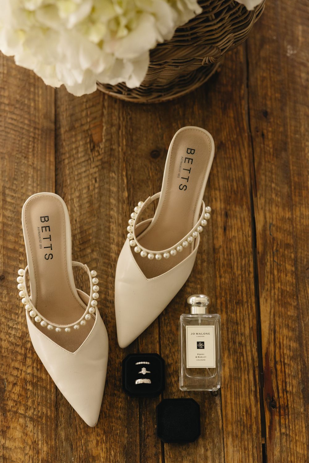 Elegant cream-colored pearl-trimmed mules with pointed toes displayed next to perfume bottles on rustic wooden surface.