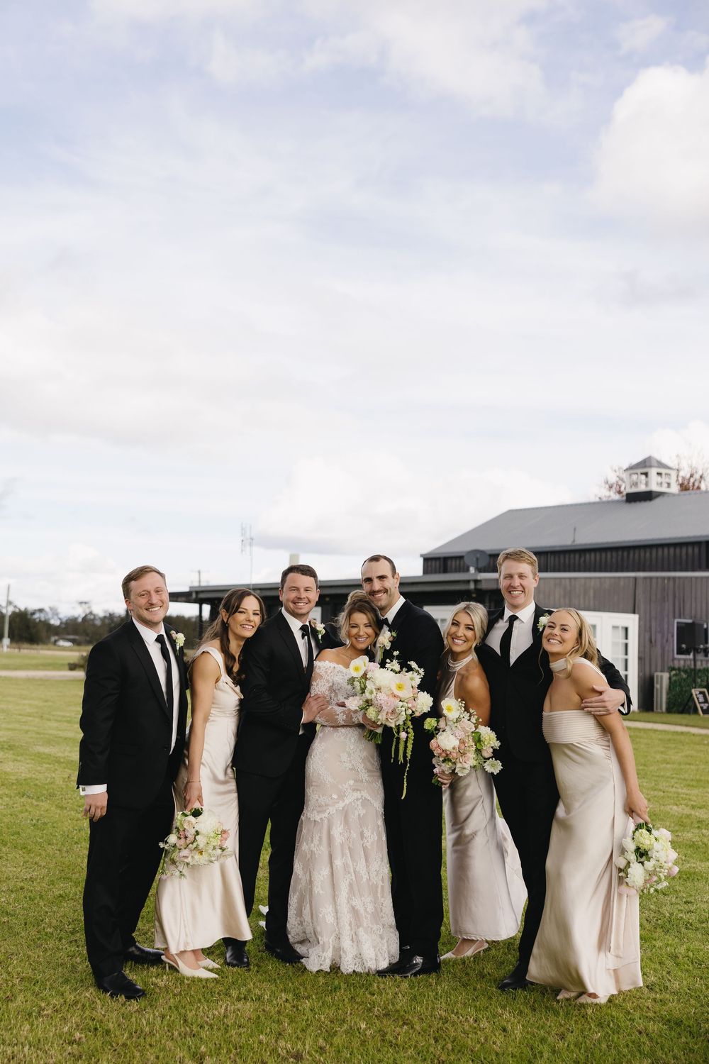 Wedding party in formal attire poses together on green lawn with rustic barn venue in background.