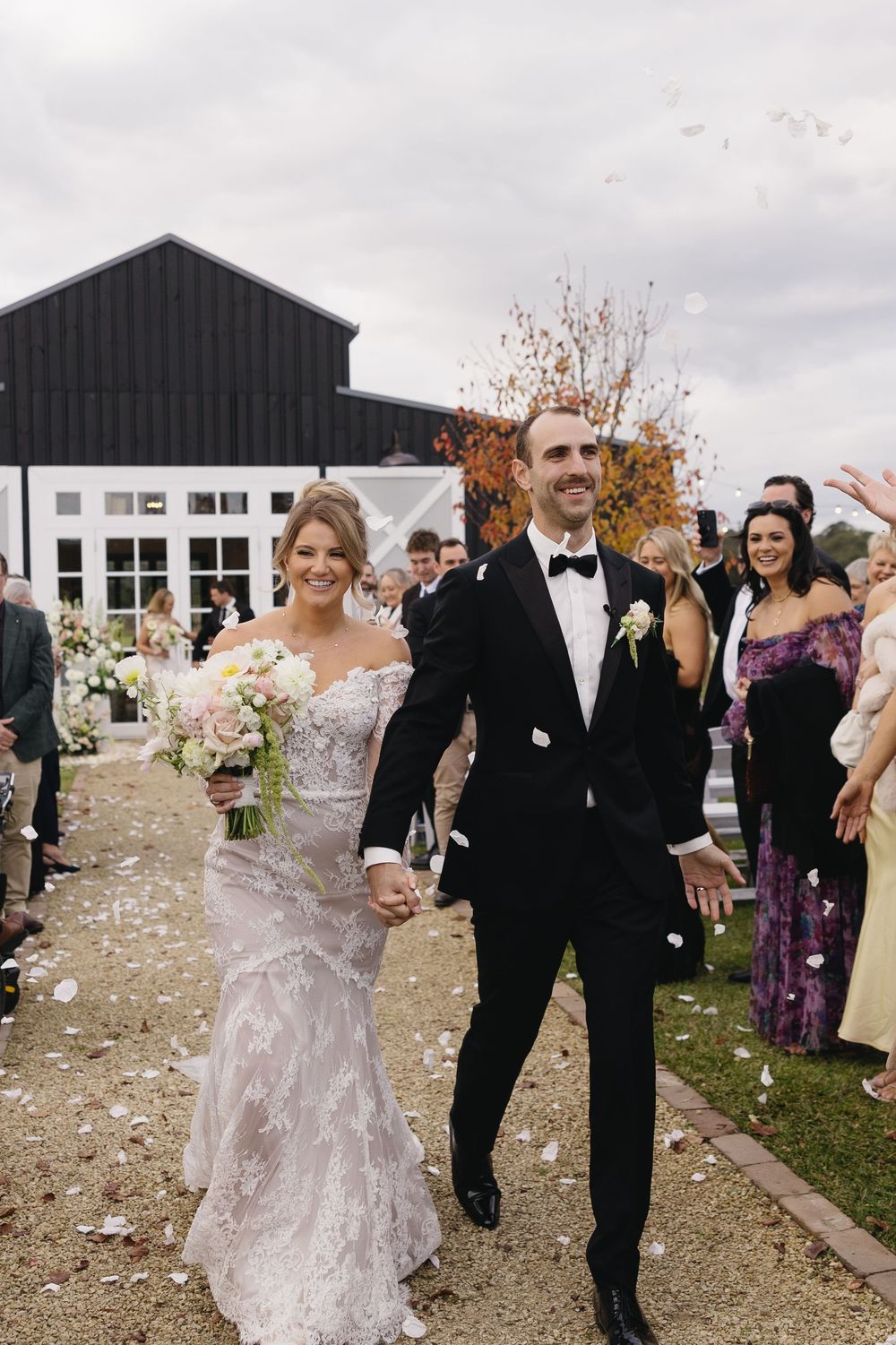 Newlywed couple walks down pathway while guests throw petals outside black barn wedding venue on cloudy day.