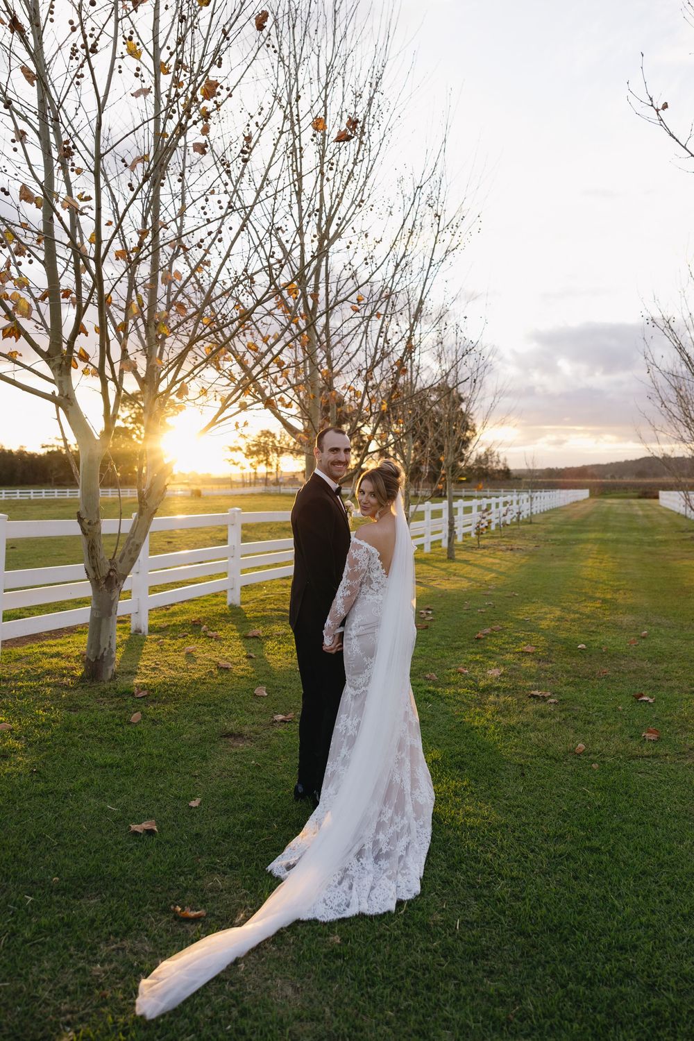 A couple in wedding attire shares an intimate moment at sunset near a white fence with trees in the background.
