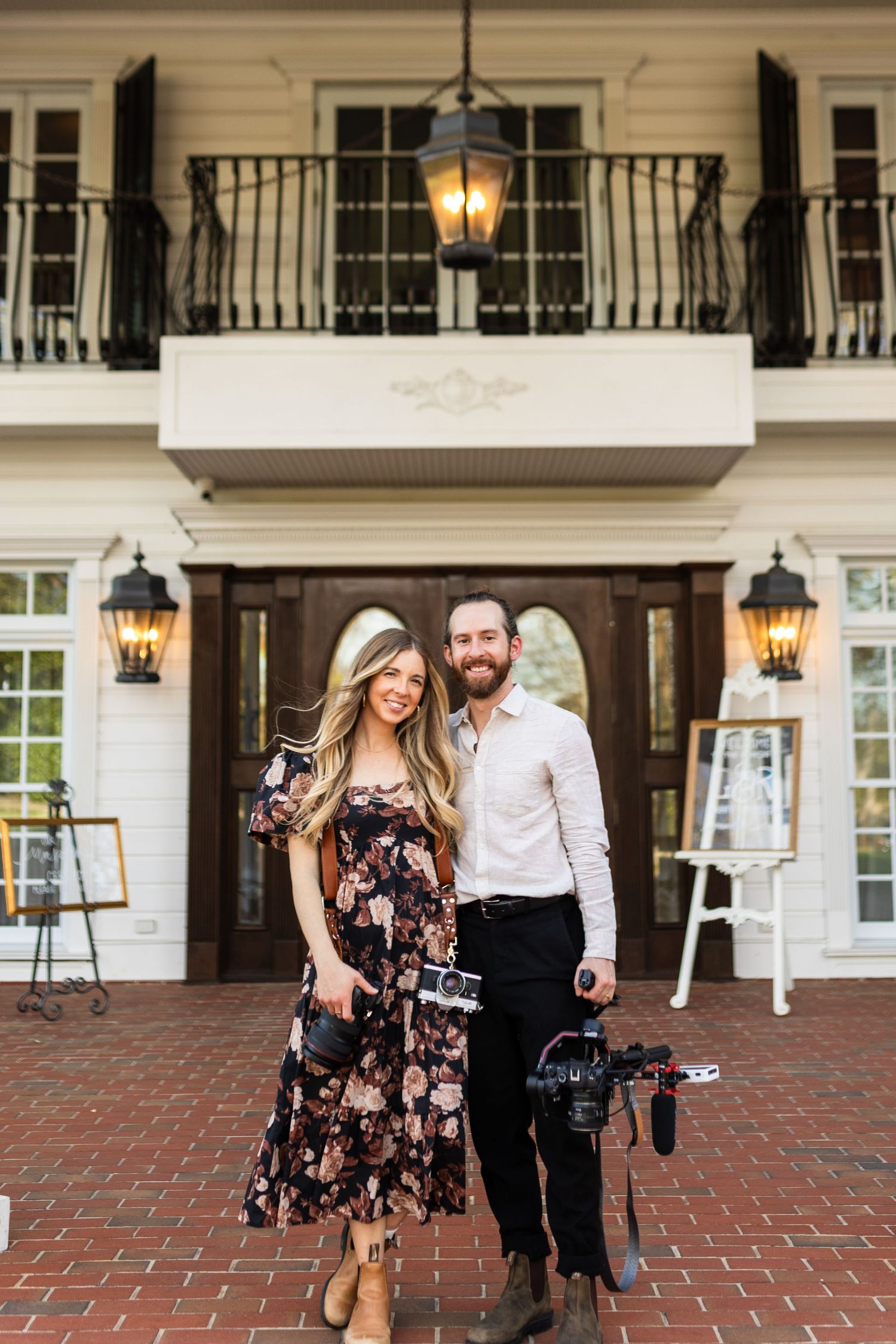 A couple stands outside a grand colonial-style house with white columns and ornate gas lanterns at dusk.