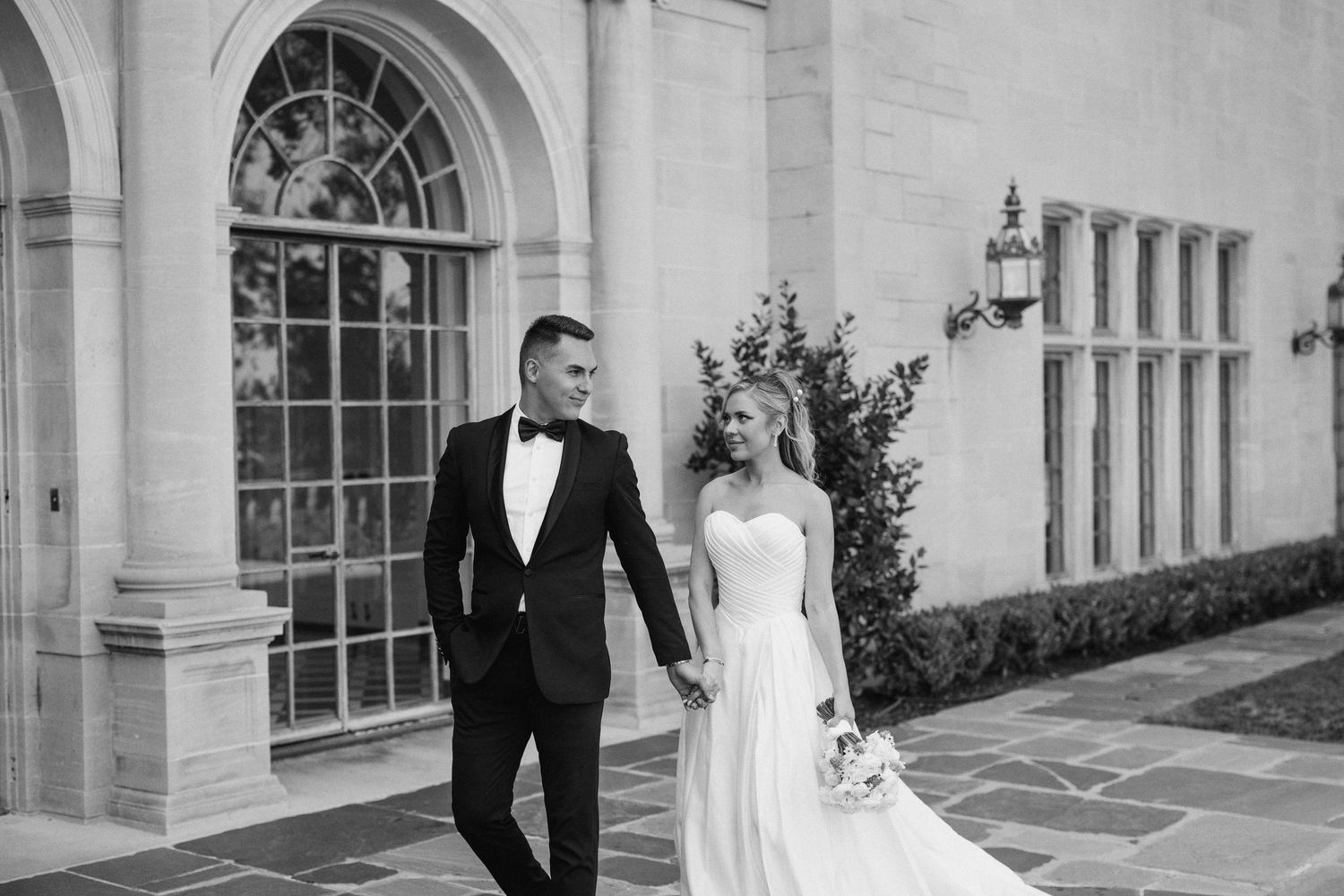 A bride and groom pose together near elegant white stone columns and arched windows of a classic building exterior.