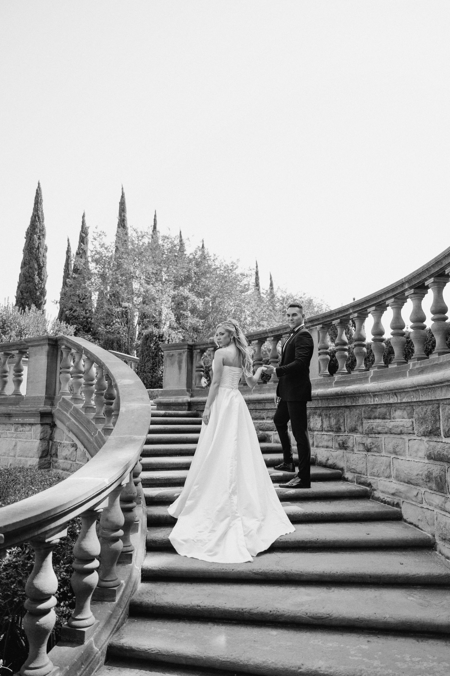 Four black and white wedding photos show a couple on grand stone steps with ornate balustrades and cypress trees in Italy.
