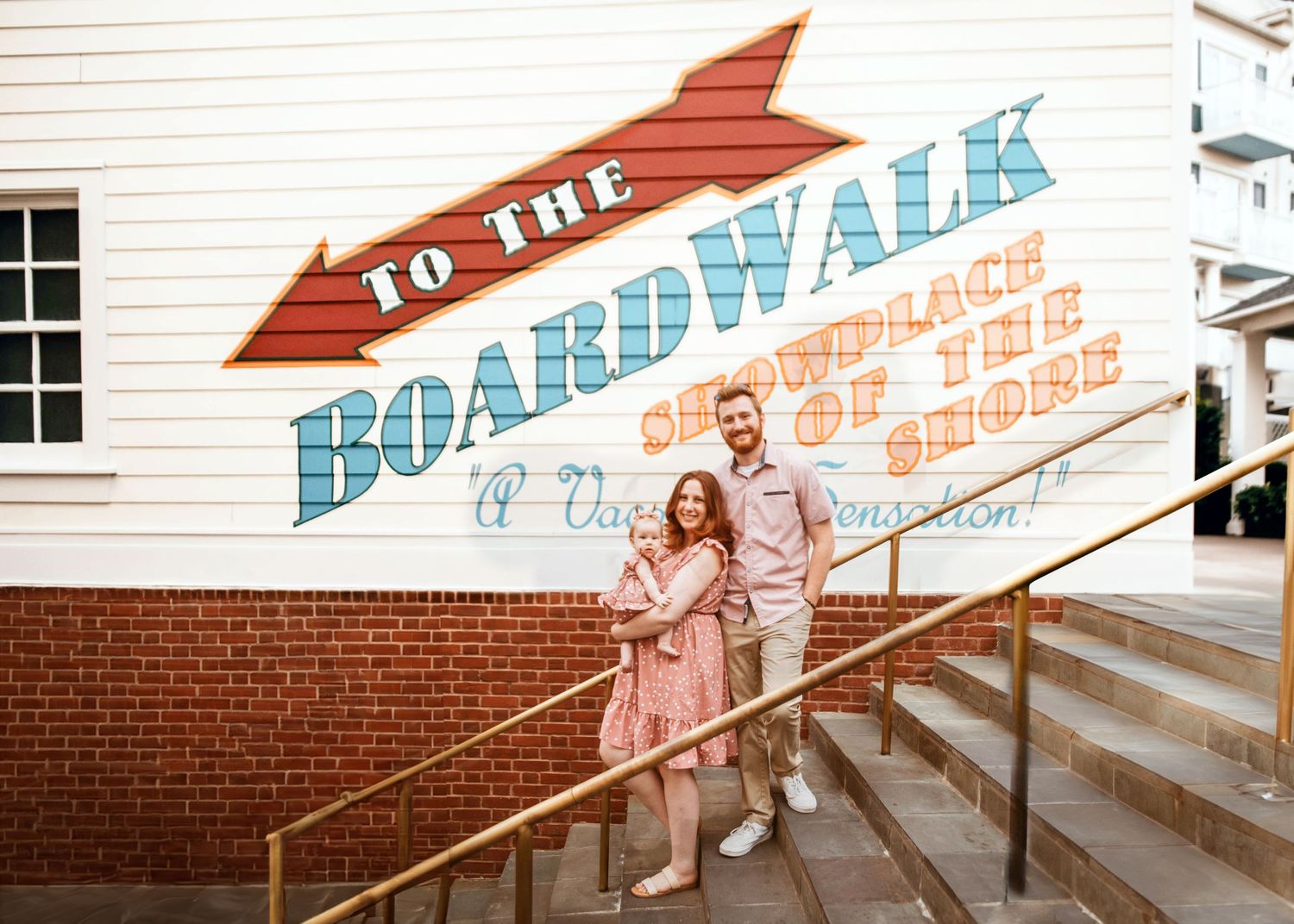 John & Emily anniversary photos at Disney Boardwalk with baby Ripley, walking down the stairs by "To The Boardwalk" mural. Cred: LexTakePix