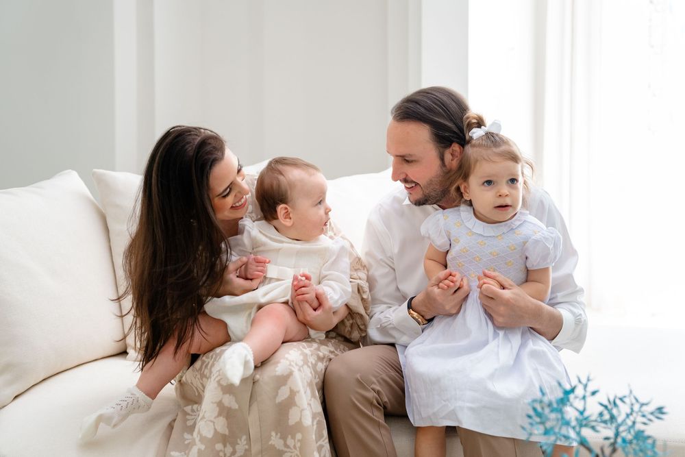 A happy family sits together on a white couch, sharing intimate moments with their children in white outfits.