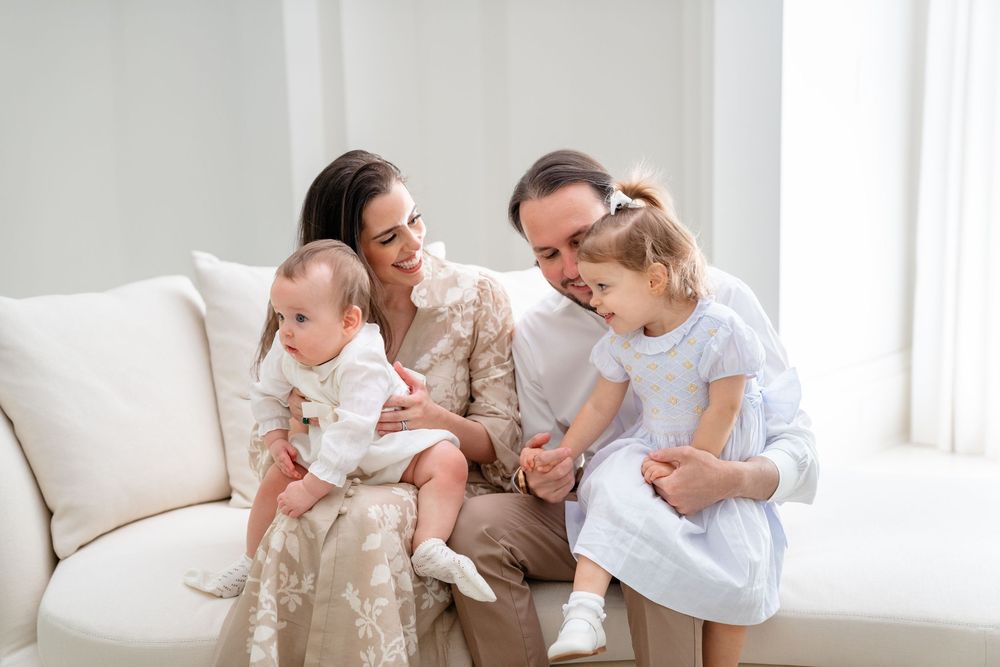 A family lounges together on a white sofa in a bright sunlit living room sharing tender moments.