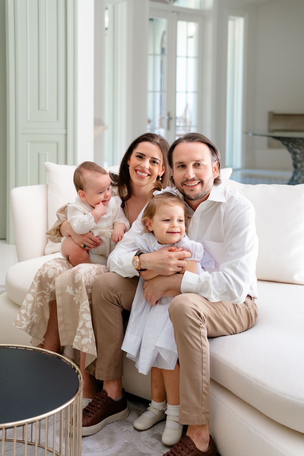 A young family in matching beige and white outfits sits together on a white sofa in a bright, modern living room.
